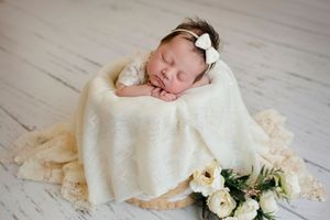 Newborn baby sleeping peacefully in a cream wrap with a white bow headband beside a bouquet of white flowers.