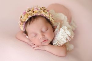 Sleeping newborn baby wearing a delicate floral crown on a soft pink backdrop.