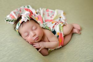 Newborn baby girl sleeping peacefully in floral wrap and matching bow headband on green backdrop.