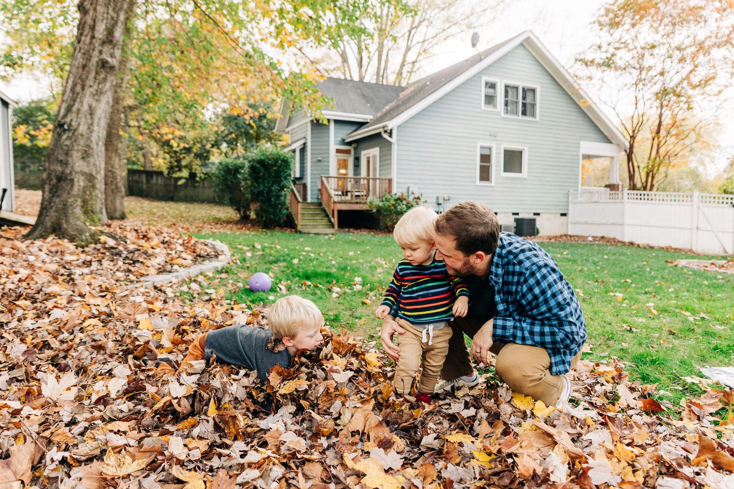 Playing in a pile of autumn leaves in front of a gray house with a deck on a fall day.