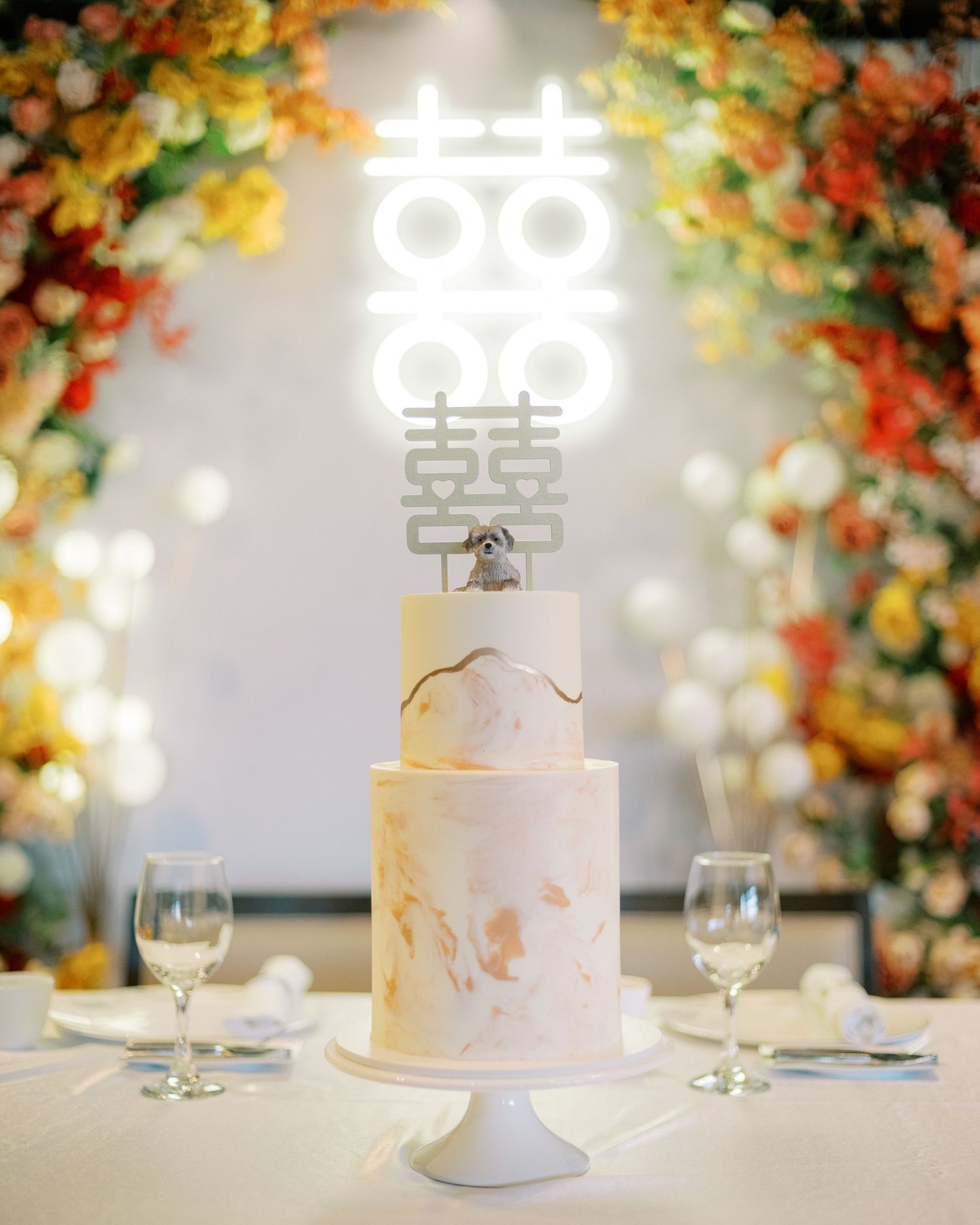 White wedding cake with Chinese double happiness symbol topper surrounded by autumn-colored floral arch.