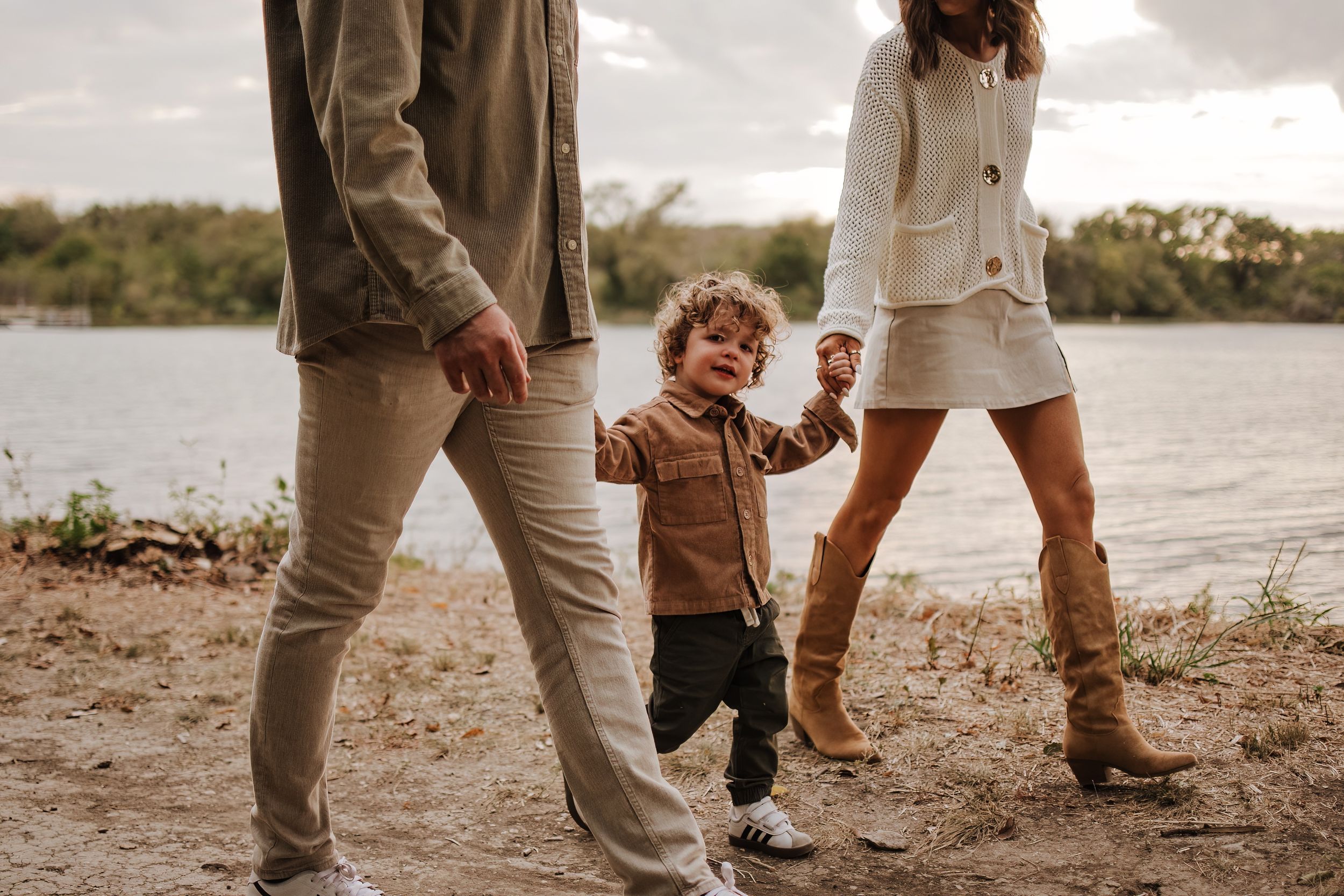 A young child walks between two people along a lakeside trail in autumn.