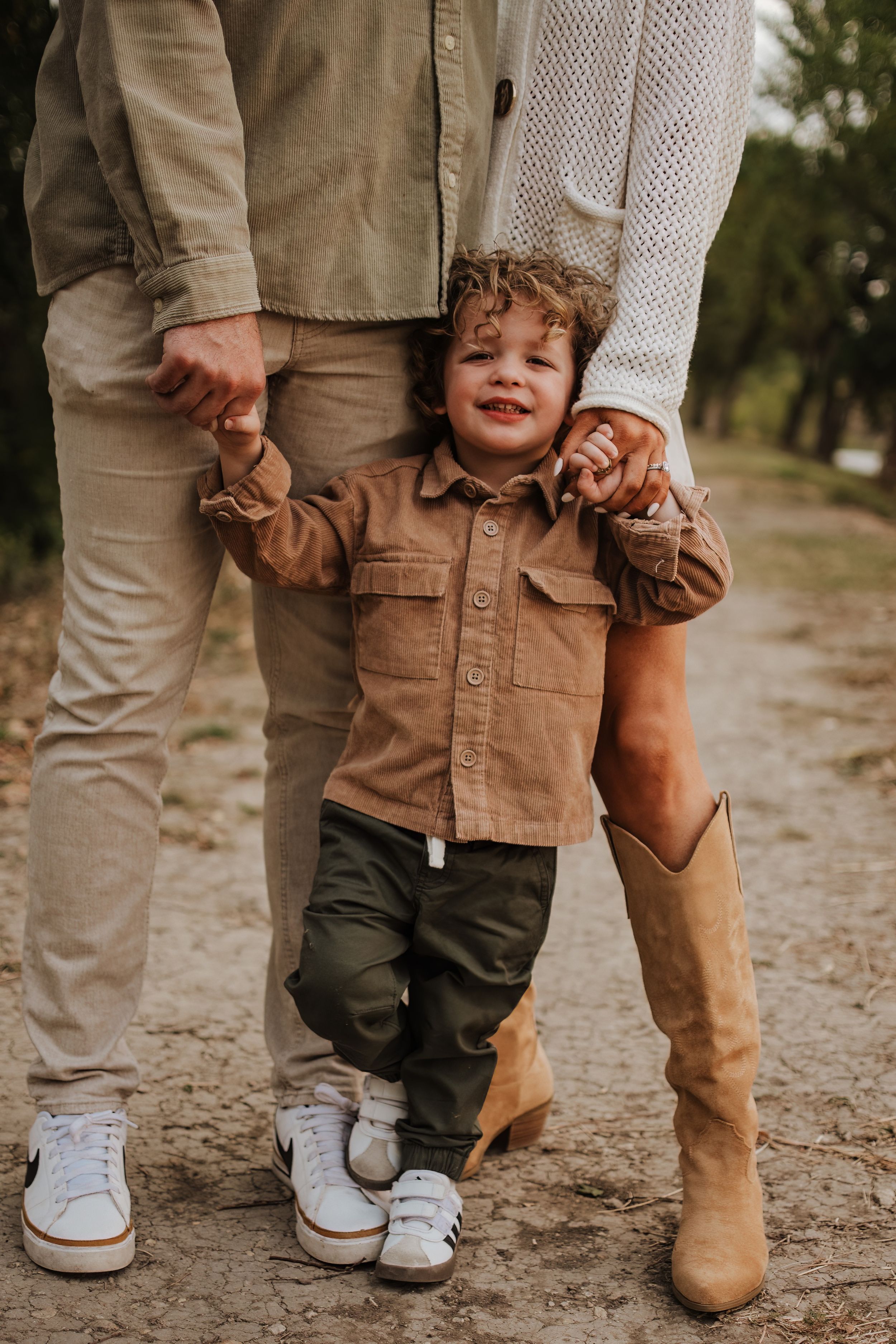 A toddler in brown clothing holds hands with adults while walking on a dirt path.