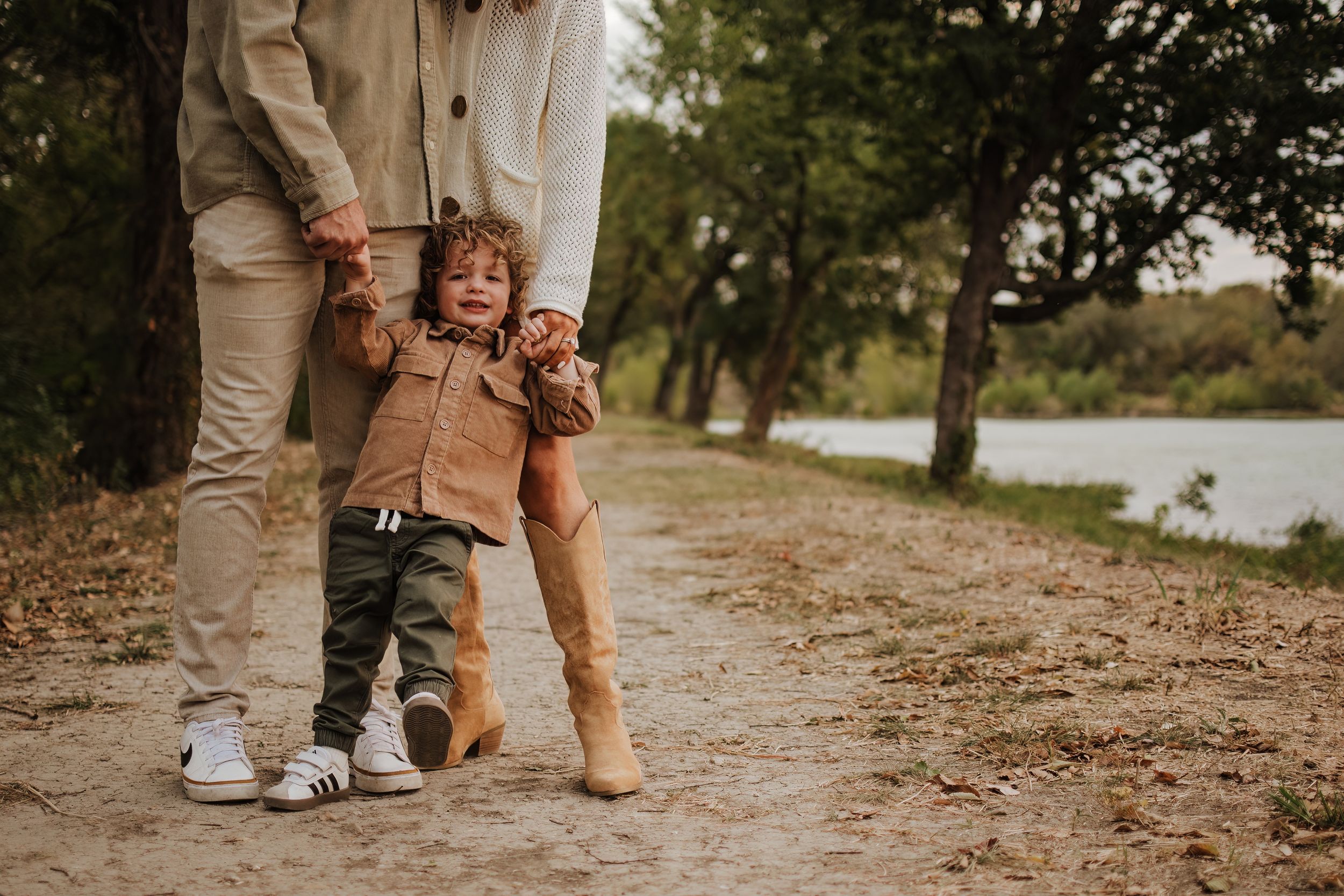 A toddler in a brown jacket takes first steps on a nature path while being supported by adults.