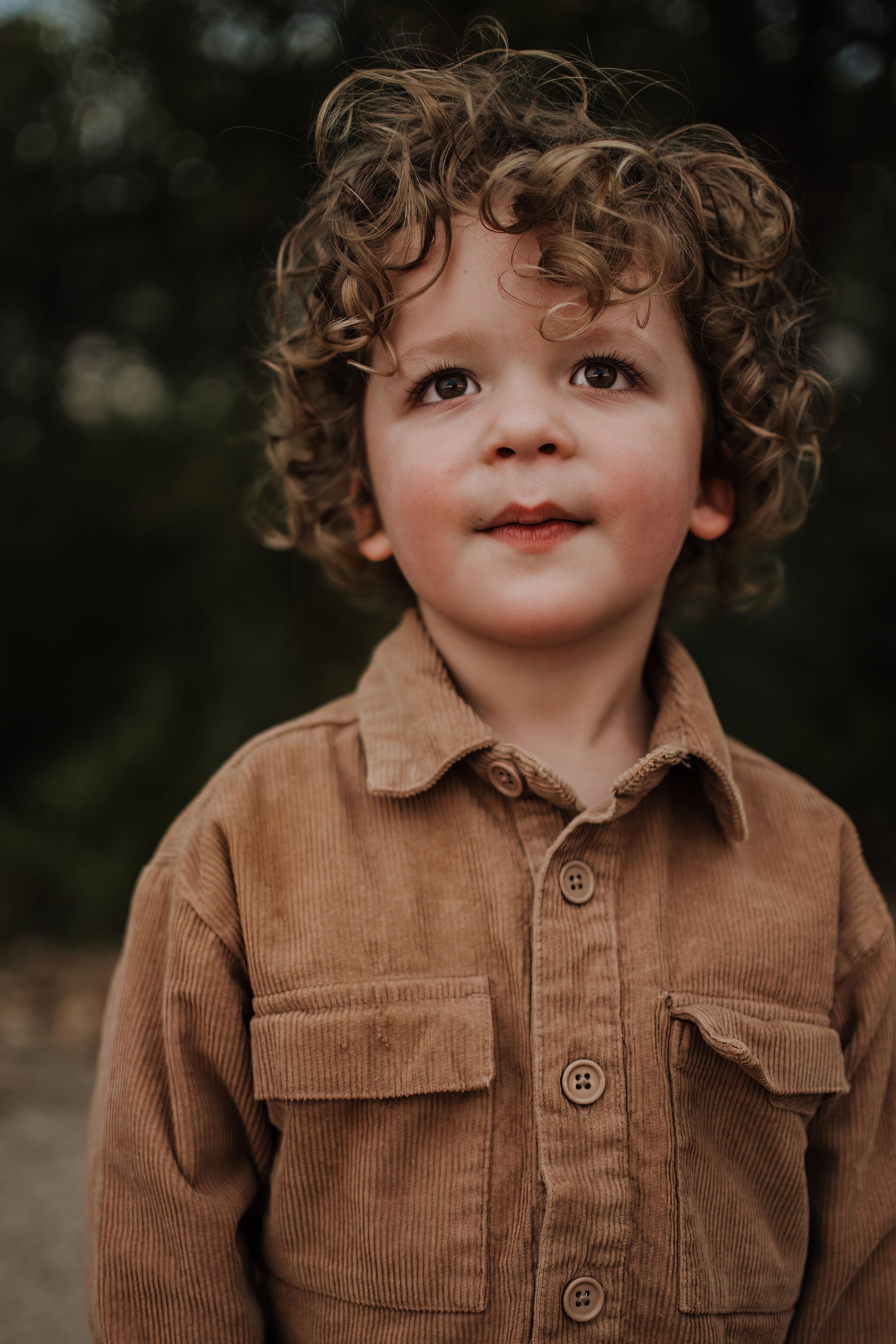 Young child in corduroy jacket smiles sweetly at camera during outdoor photo session.
