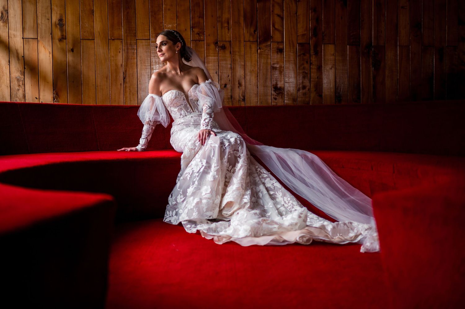 Wedding dress displayed dramatically on red carpet in rustic wooden interior.
