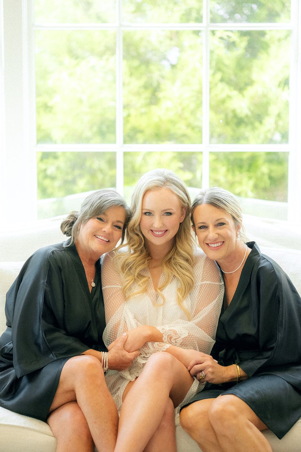 Three women sit together on a couch, with the middle figure in white and the others wearing black robes.