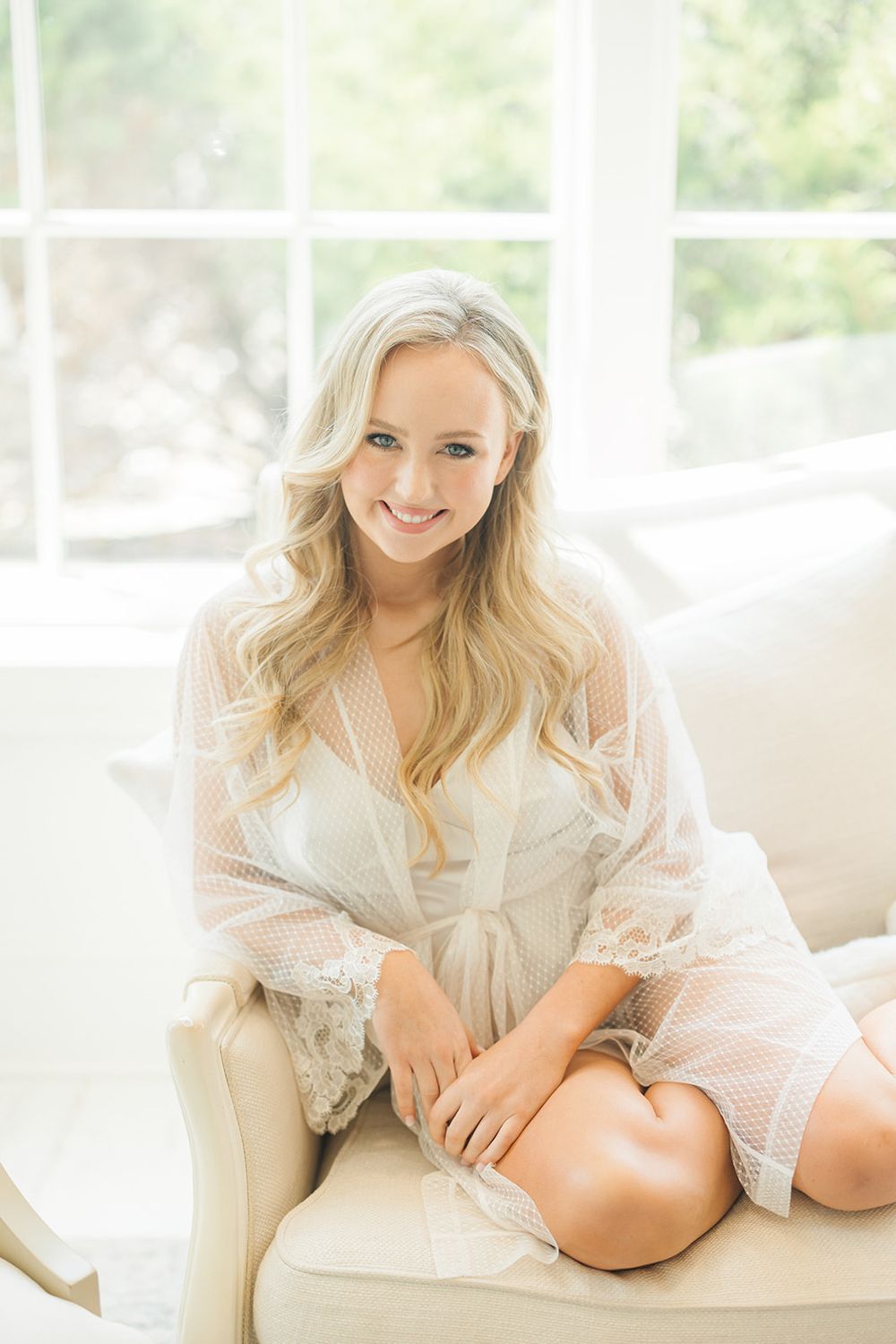 A blonde woman in a white lace robe sits on a cream-colored couch near a bright window.