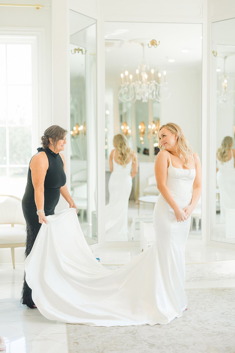 Two people in formal attire prepare a wedding dress train in an elegant white room with mirrored walls.