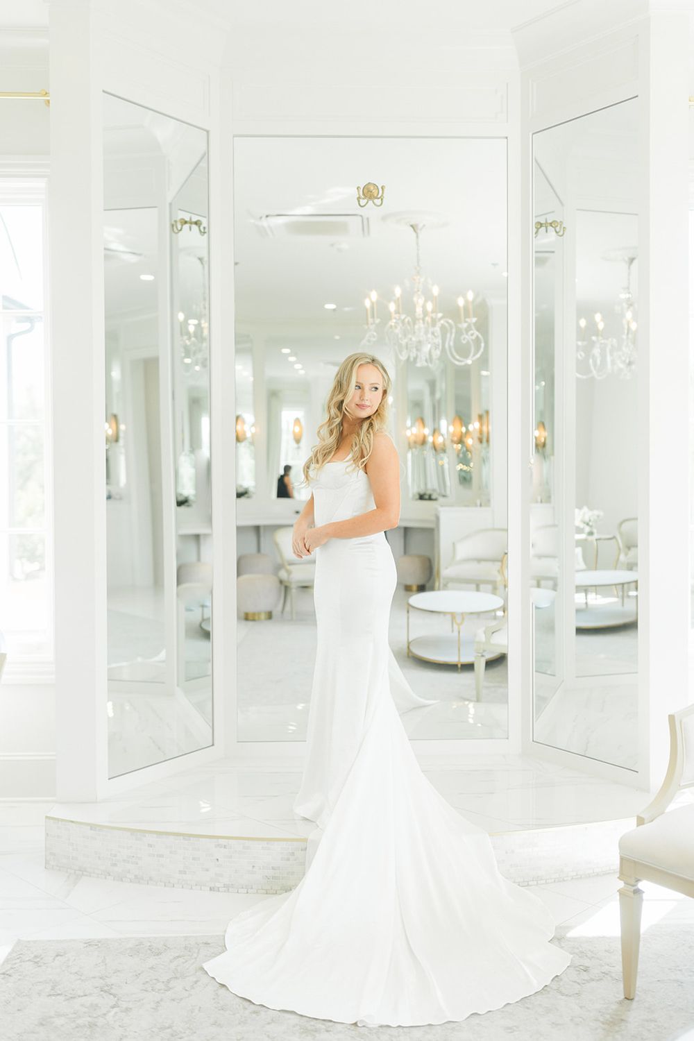 A bride in a white wedding gown poses elegantly in a luxurious all-white room with crystal chandeliers and mirrors.