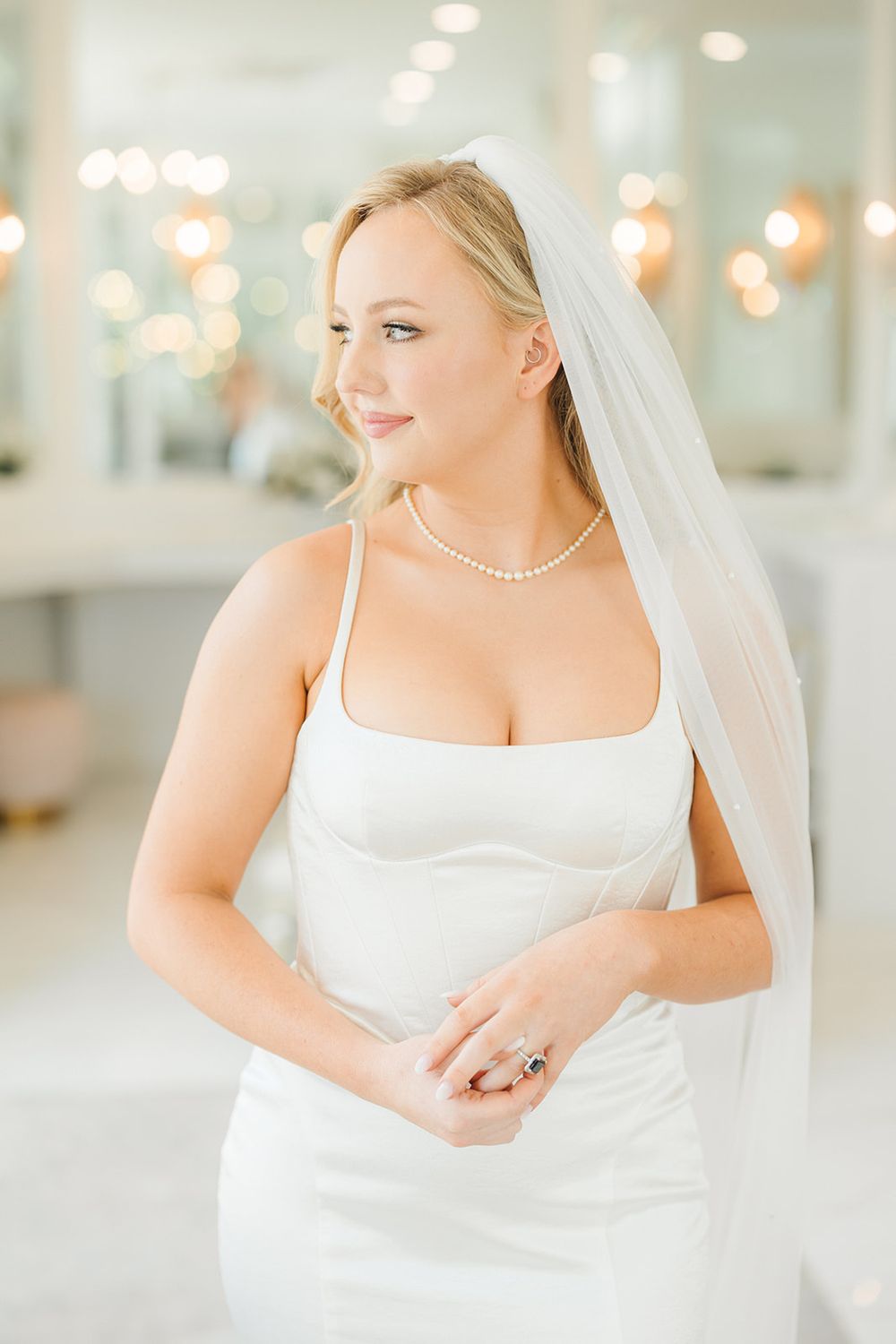 Beautiful bride in elegant white wedding gown with pearl necklace and flowing veil poses in bright indoor setting.