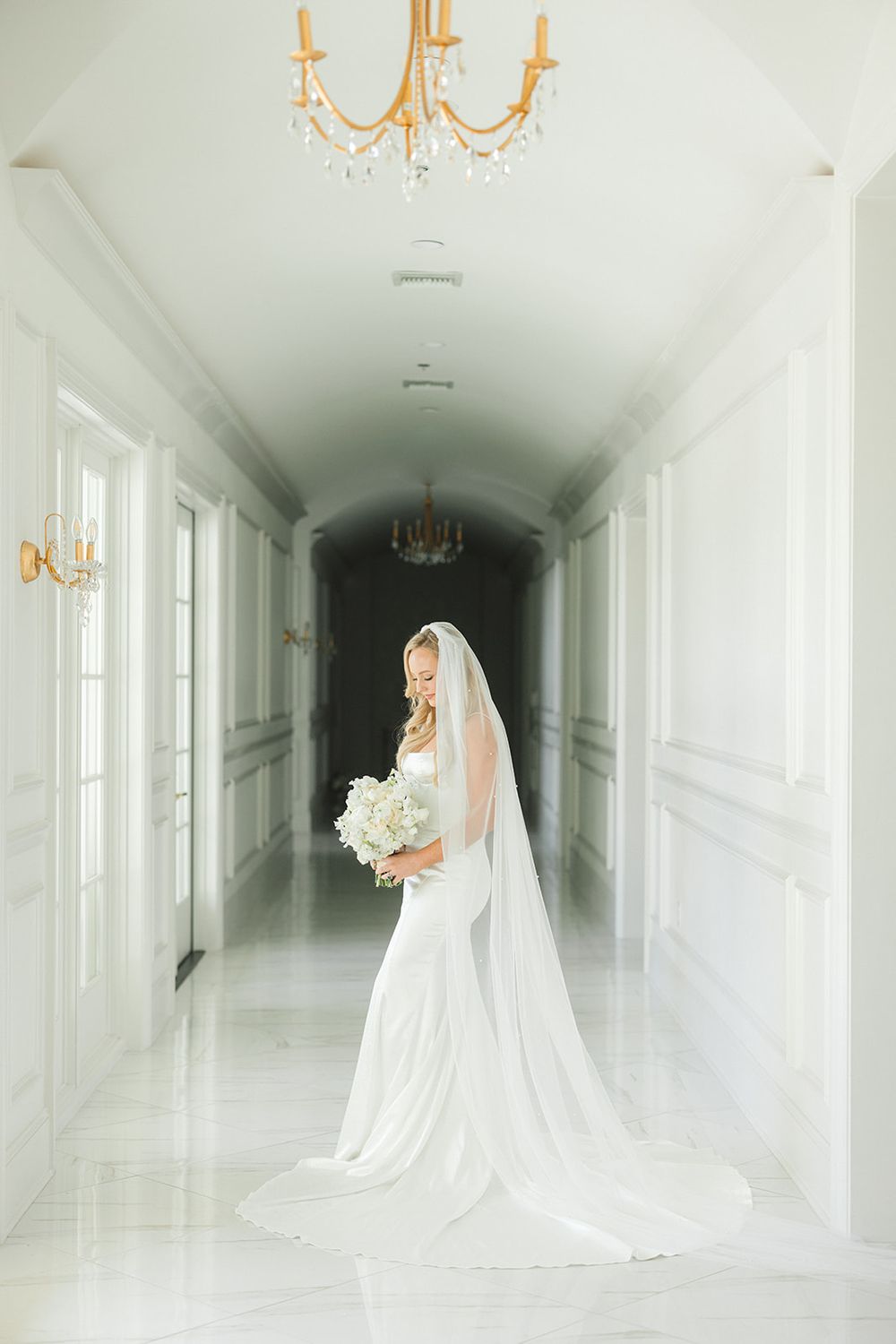 A flowing white wedding gown and veil photographed in an elegant hallway with gold chandeliers and natural light.
