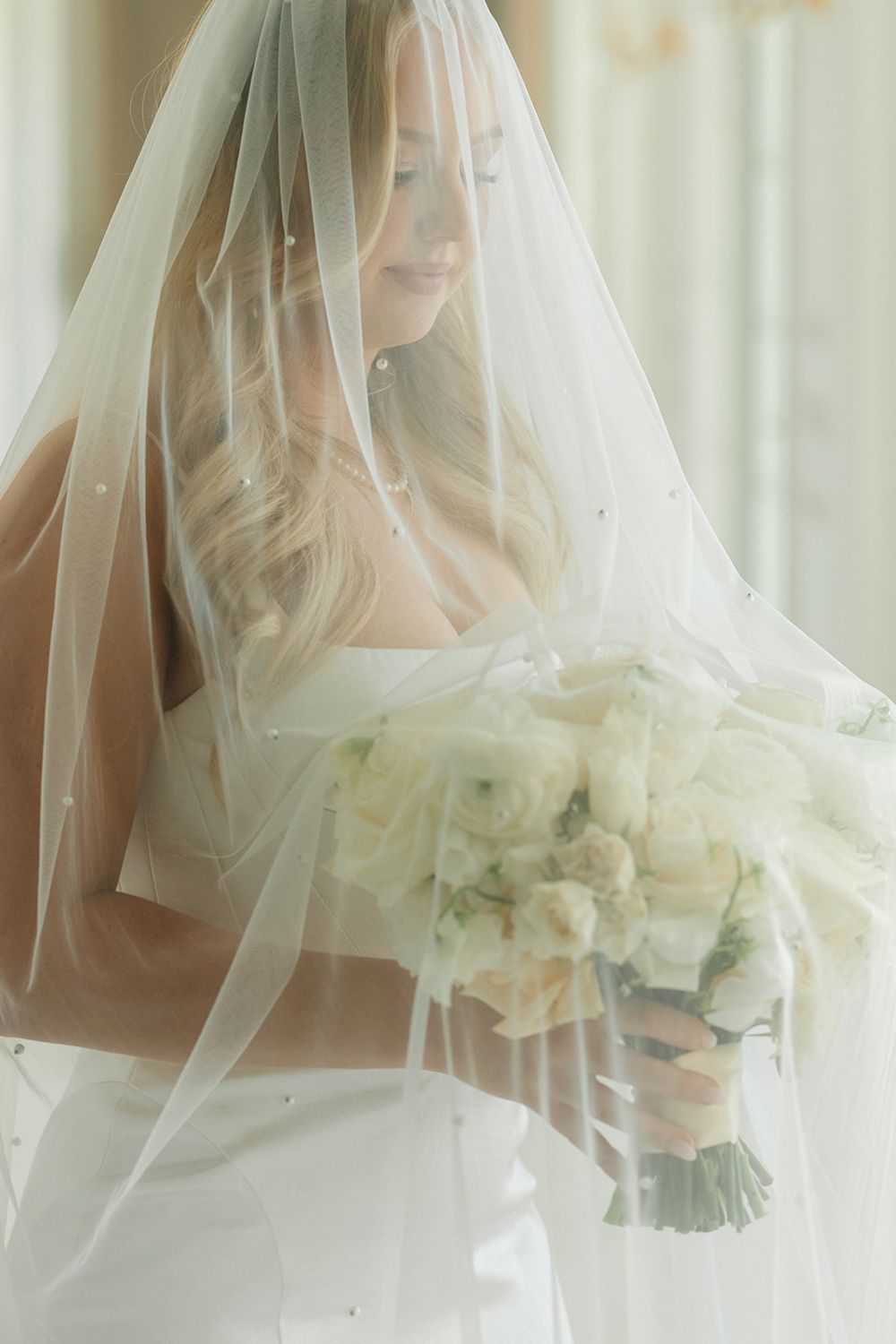 Ethereal bridal portrait with delicate white veil and ivory bouquet in soft window light.