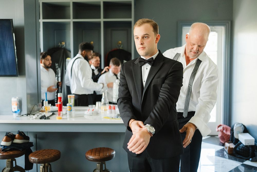 Men in formal attire prepare for an event in a room with a bar counter and wooden stools.