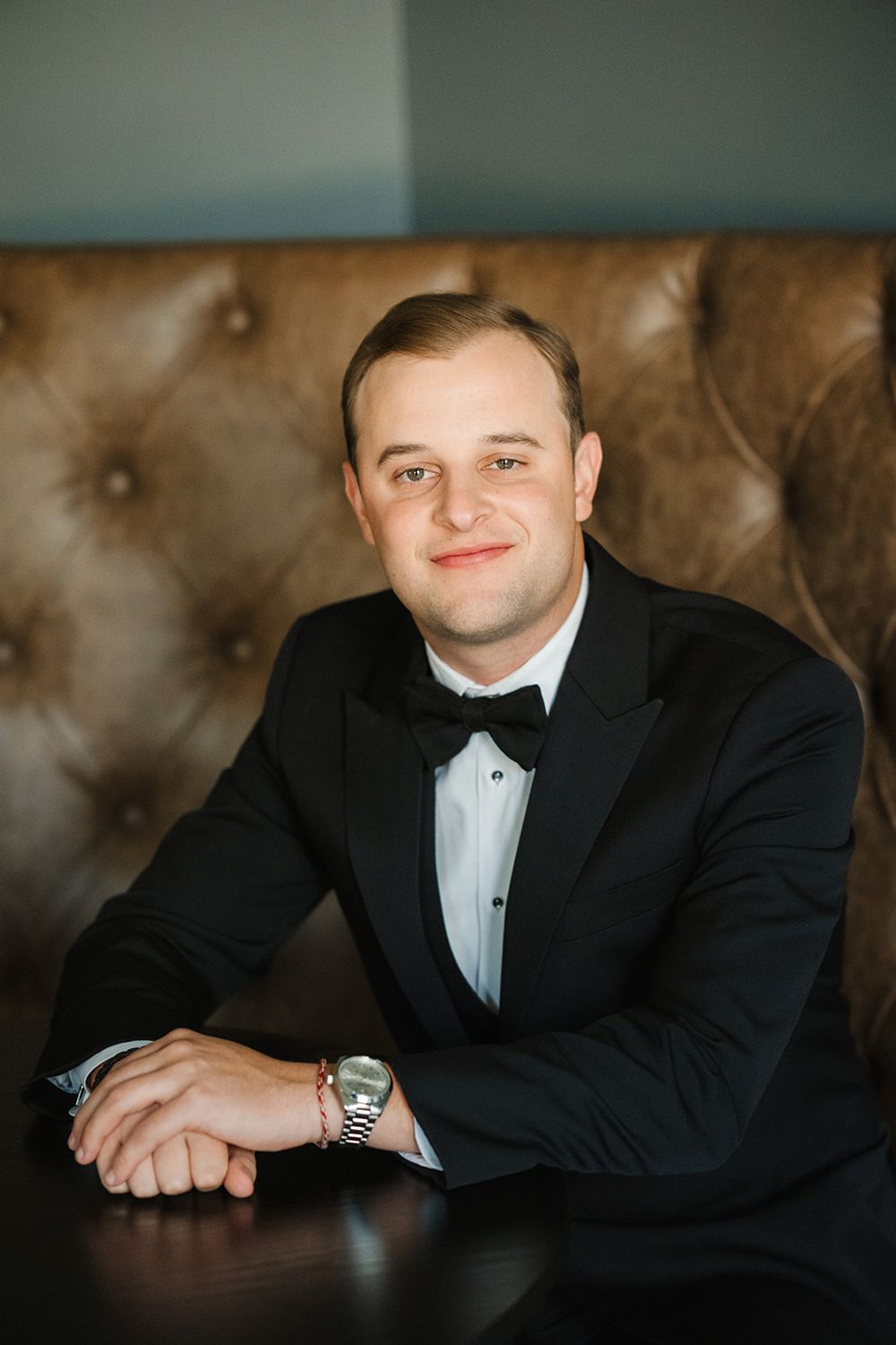 Groom in black tuxedo poses against leather upholstered background in warm lighting.