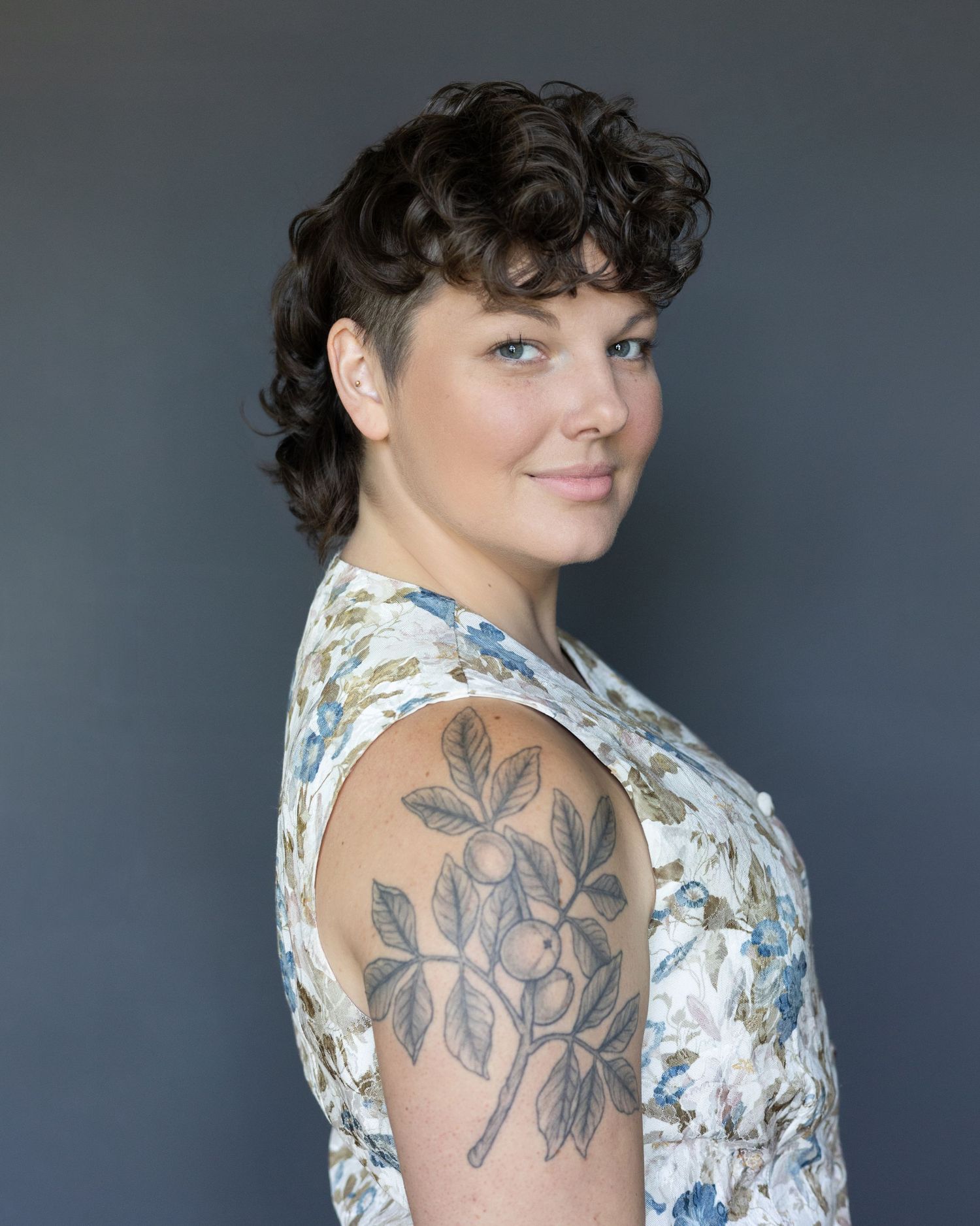 Close-up of Theo Hayes smiling softly, short hair styled, tattoos visible, against a grey backdrop, wearing a light floral vest.