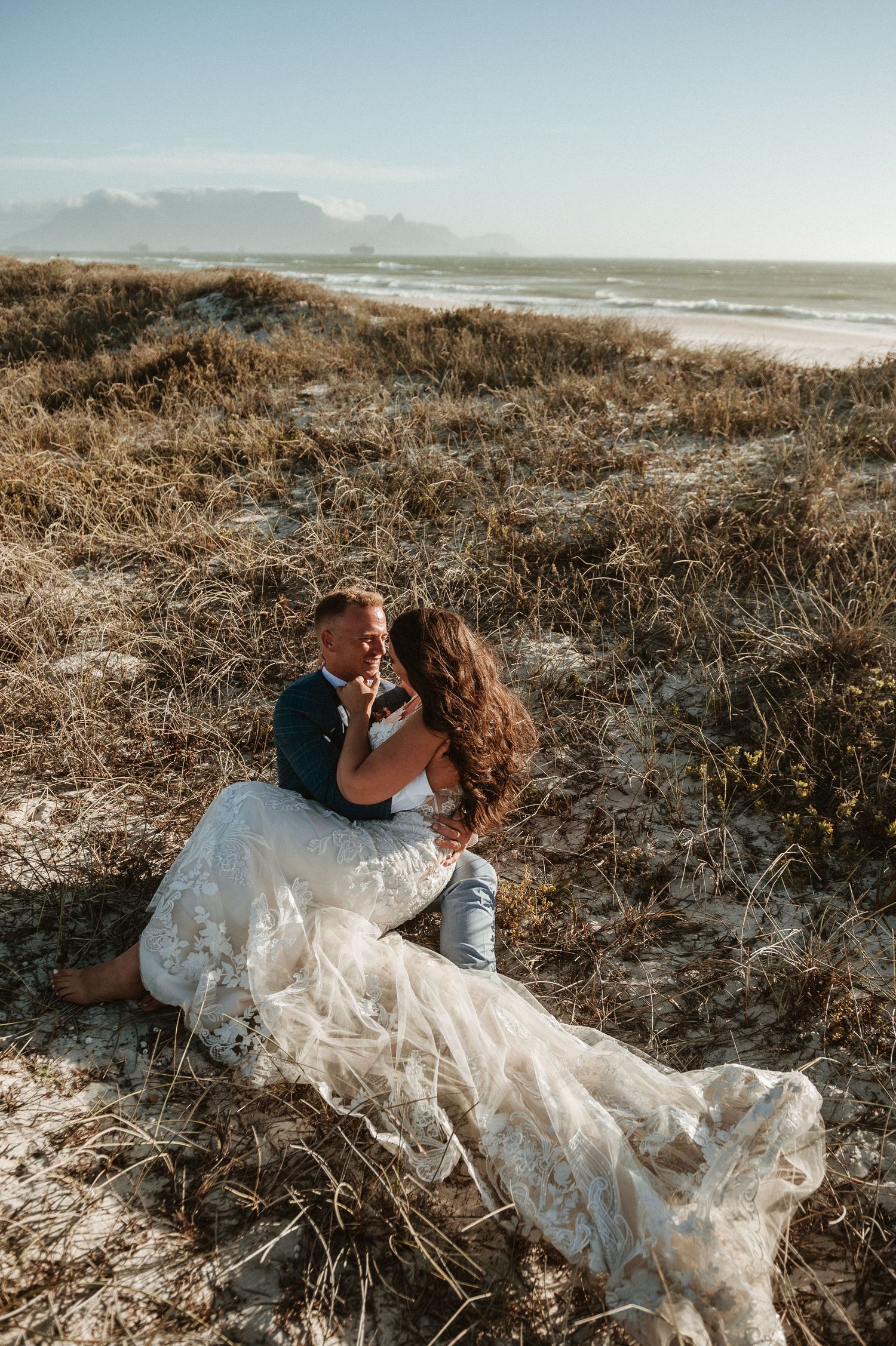 A romantic coastal wedding photoshoot with a couple sitting on a grassy cliff overlooking the ocean at sunset.