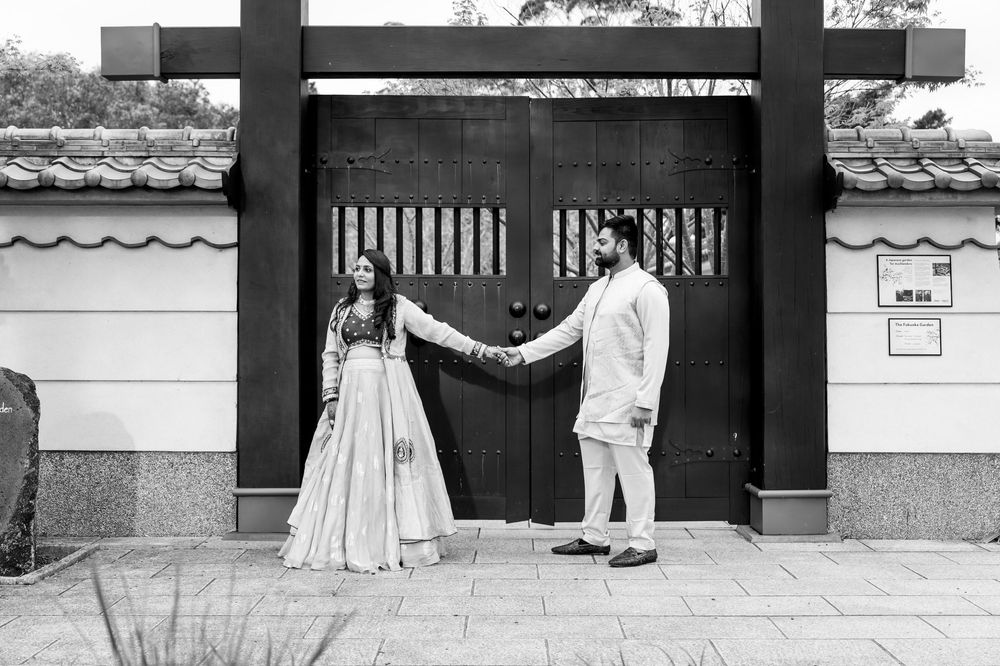 A couple in formal attire holds hands while standing in front of a traditional Asian-style gate in black and white.