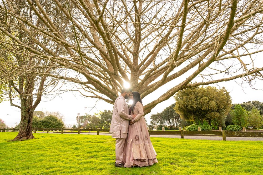 Two figures in flowing formal attire embrace under a sprawling tree on a sunny lawn.