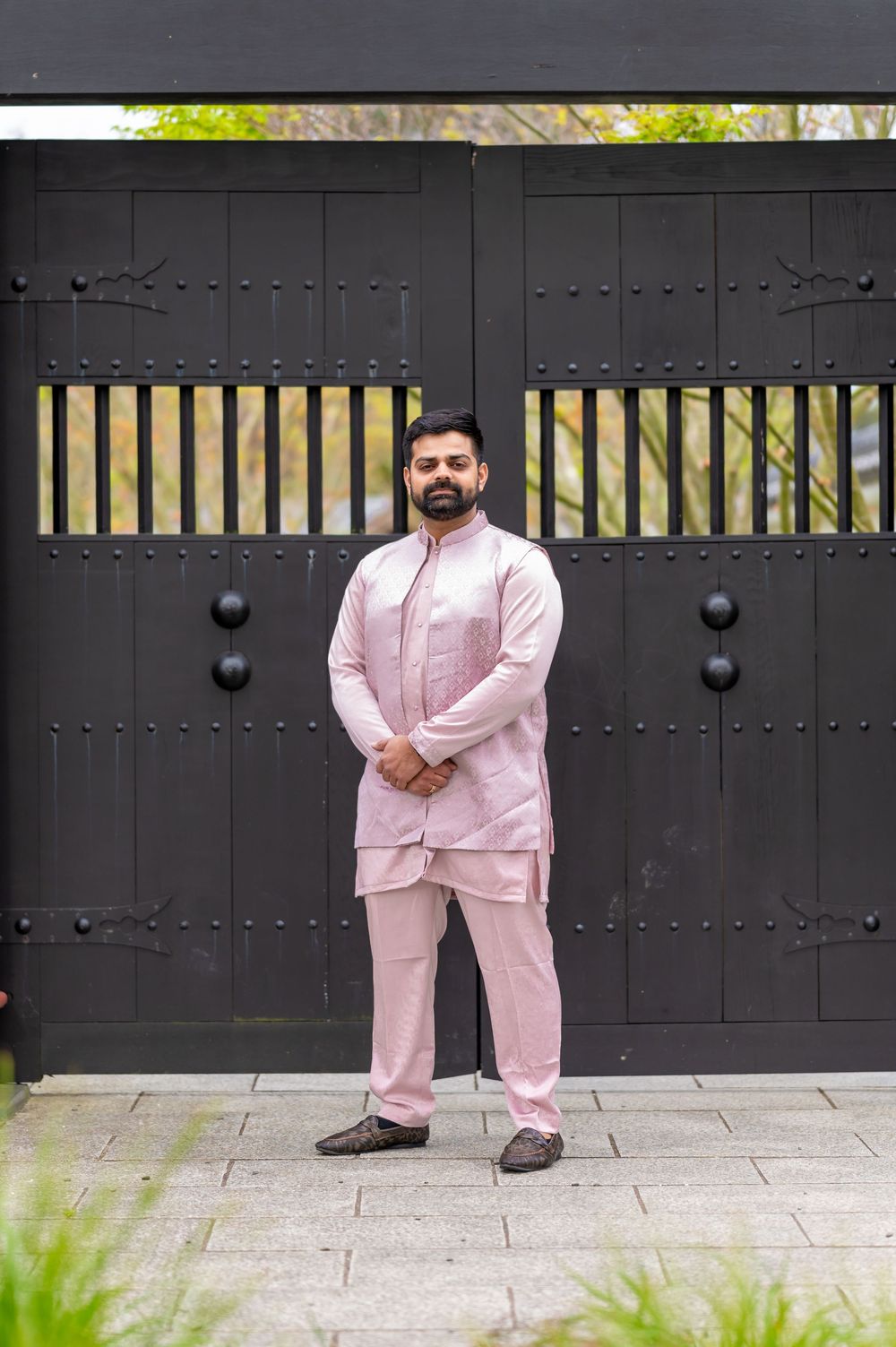 Someone stands in front of a large black gate wearing a light pink traditional kurta pajama outfit.