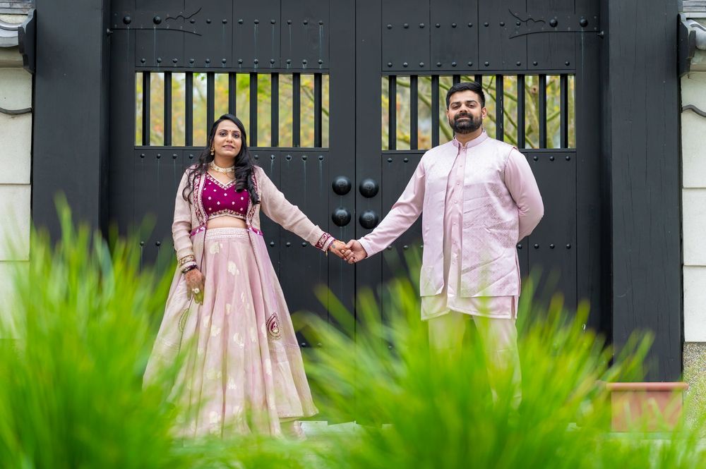 A couple in formal wedding attire stands in a garden setting with lush greenery and decorative gates.