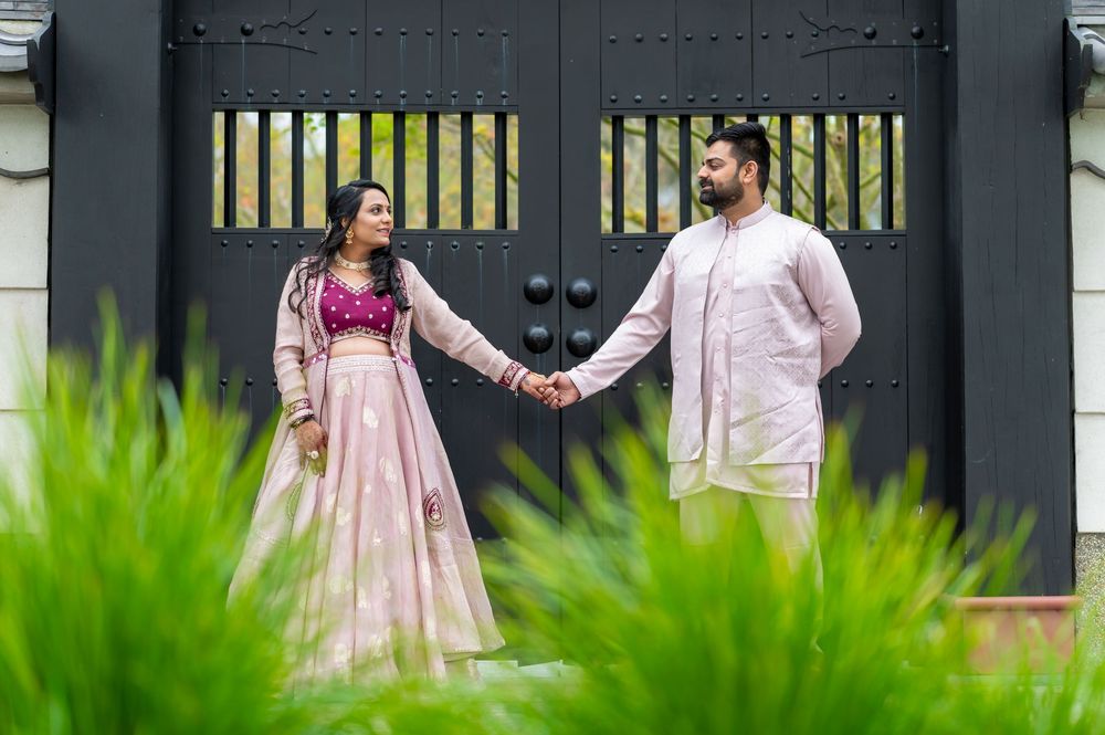 A couple in traditional Indian attire holds hands while standing in front of a black gate with green foliage in foreground.