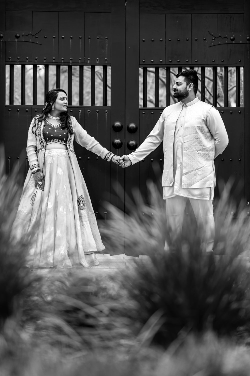 A black and white romantic couple portrait holding hands in traditional Indian attire against ornate doors with foliage.