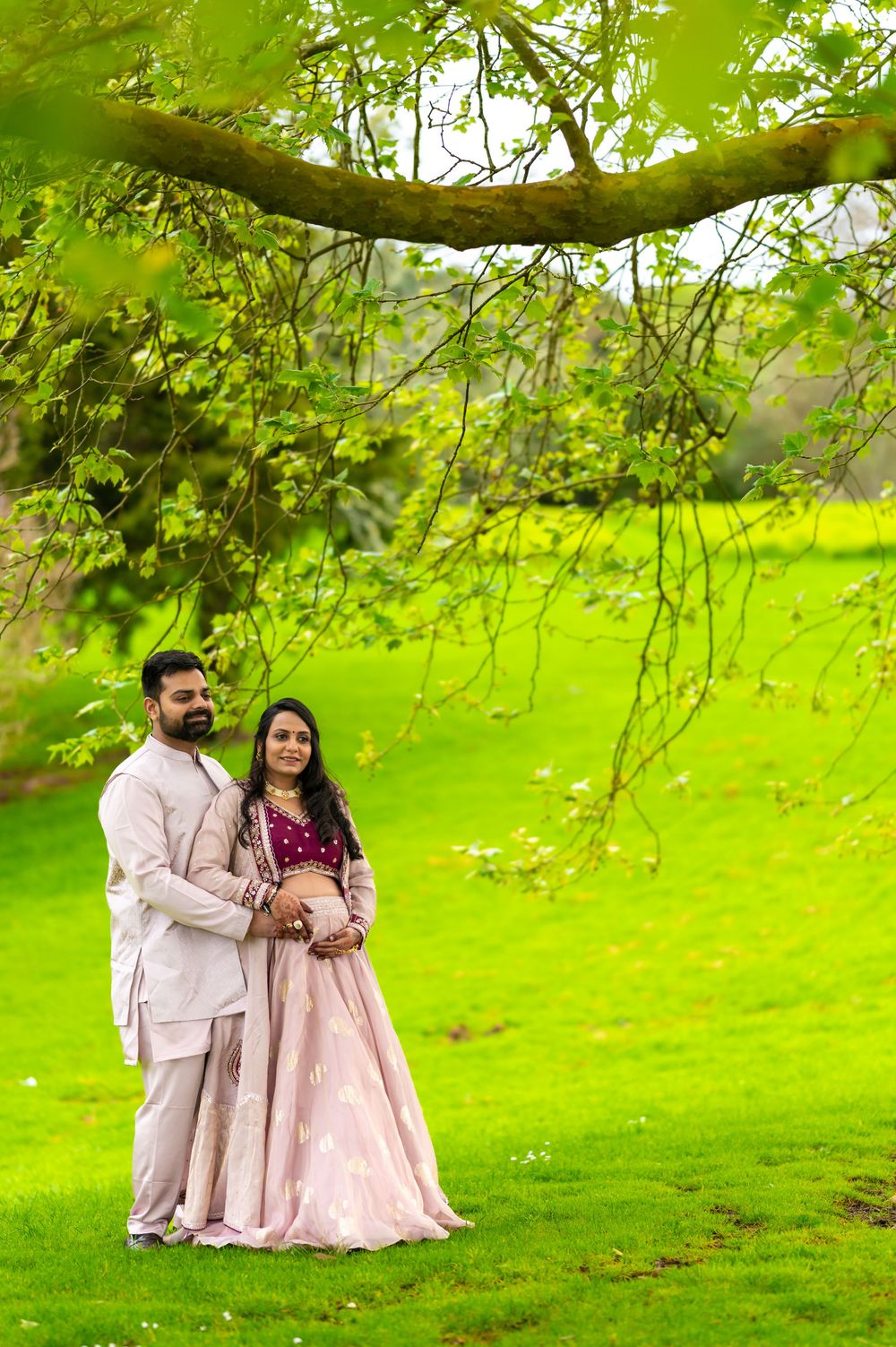 A couple in coordinating light outfits share romantic moments on a lush green lawn beneath overhanging tree branches.