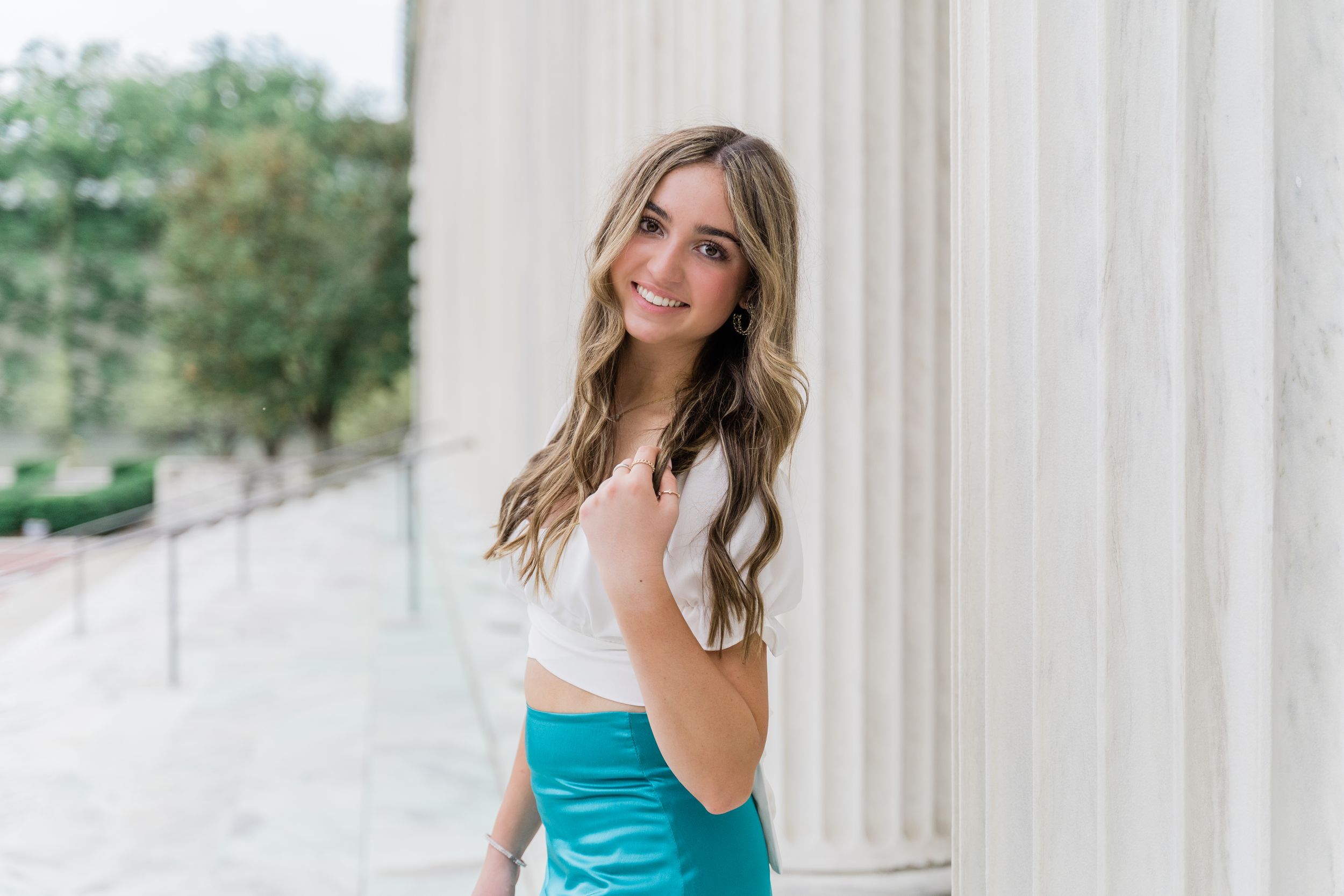 A person in a bright turquoise dress poses elegantly between white columns on a sunny day.