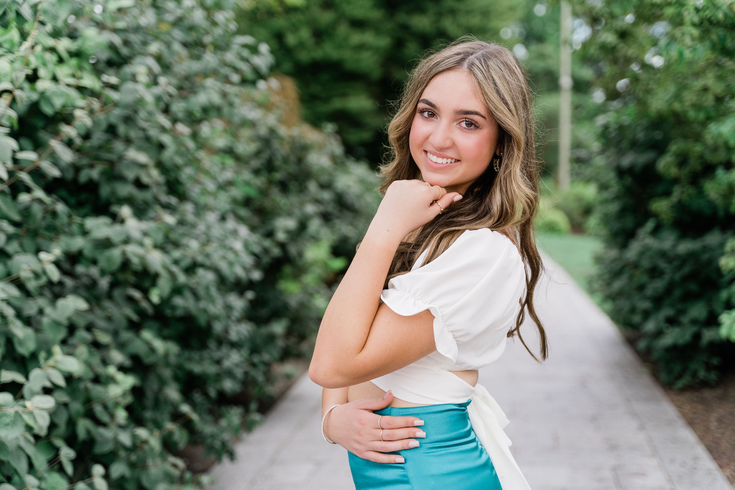 Someone wearing a white blouse and turquoise skirt poses on a garden path surrounded by greenery.