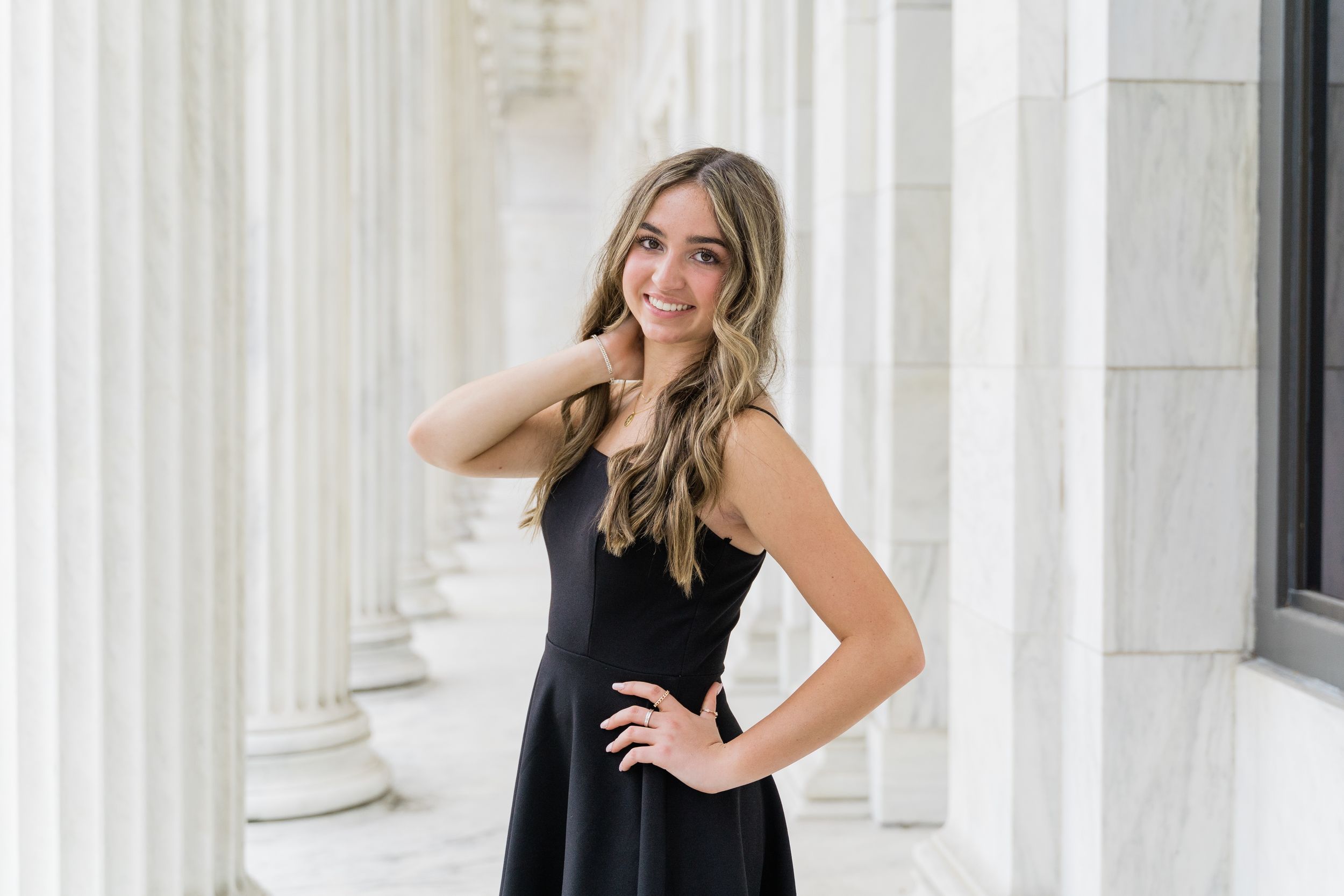 A woman in a black sleeveless dress poses between white marble columns with a bright smile.