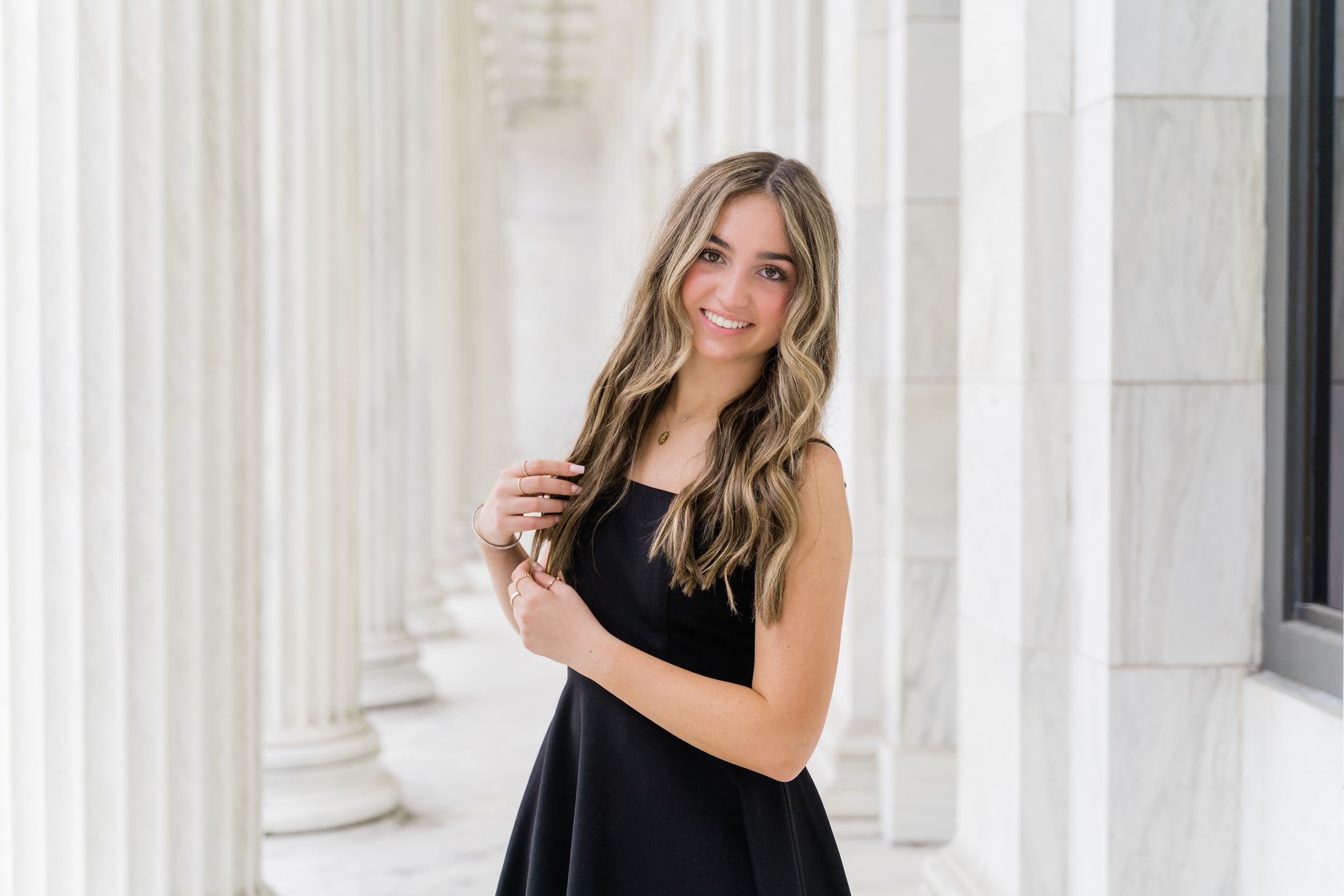 A person in a black sleeveless dress poses elegantly between white marble columns with long blonde hair.