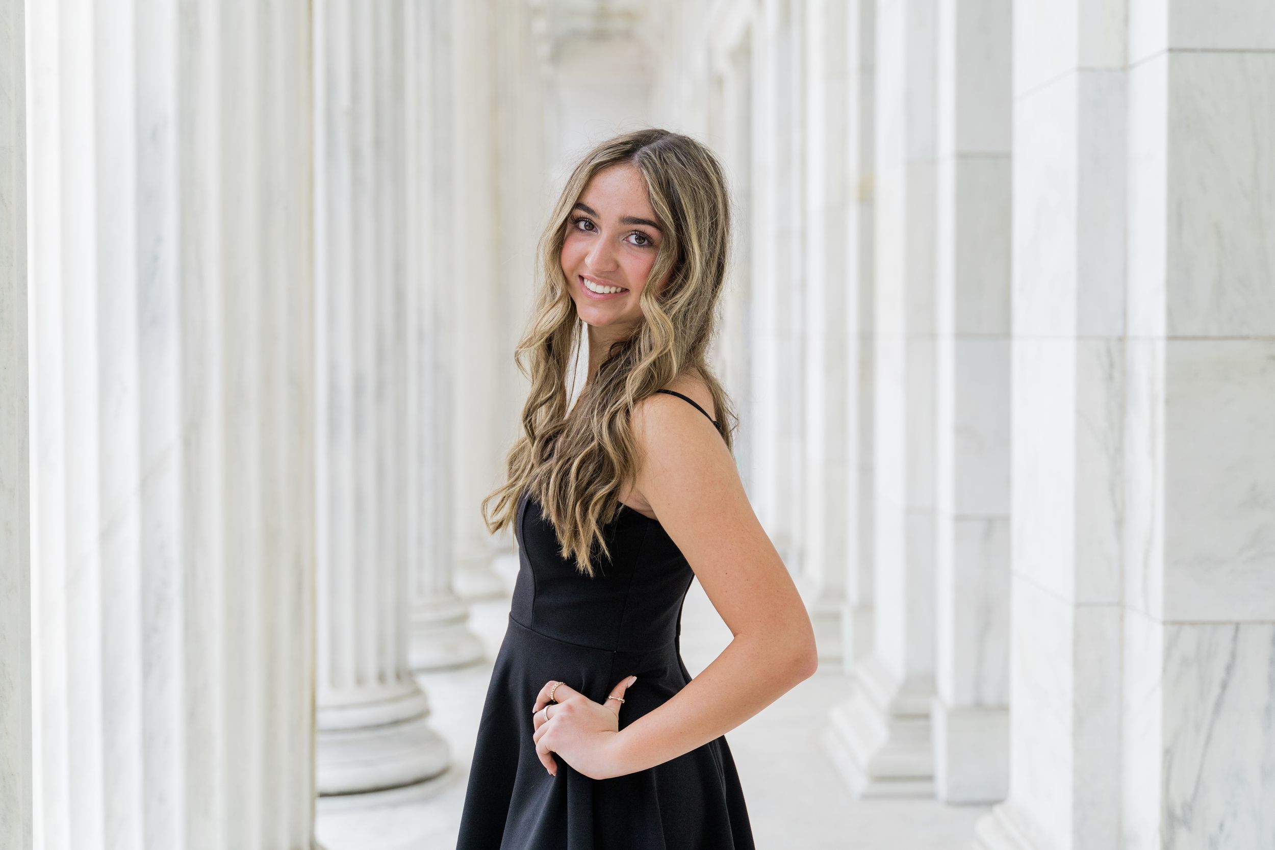 A blonde woman in an elegant black dress poses against white columns in an outdoor photoshoot setting.
