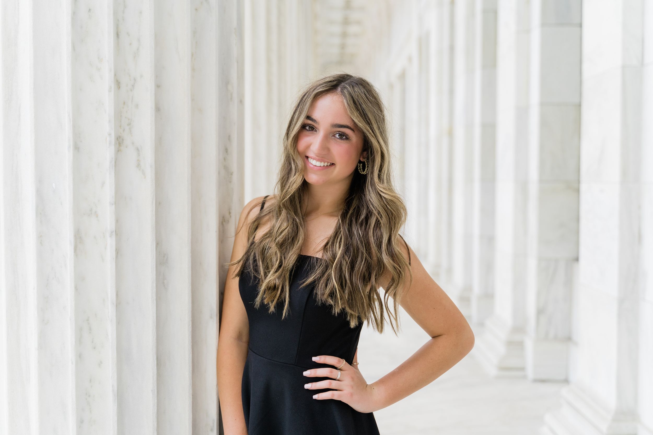 Person in a black sleeveless top with long wavy blonde hair poses against white columns.