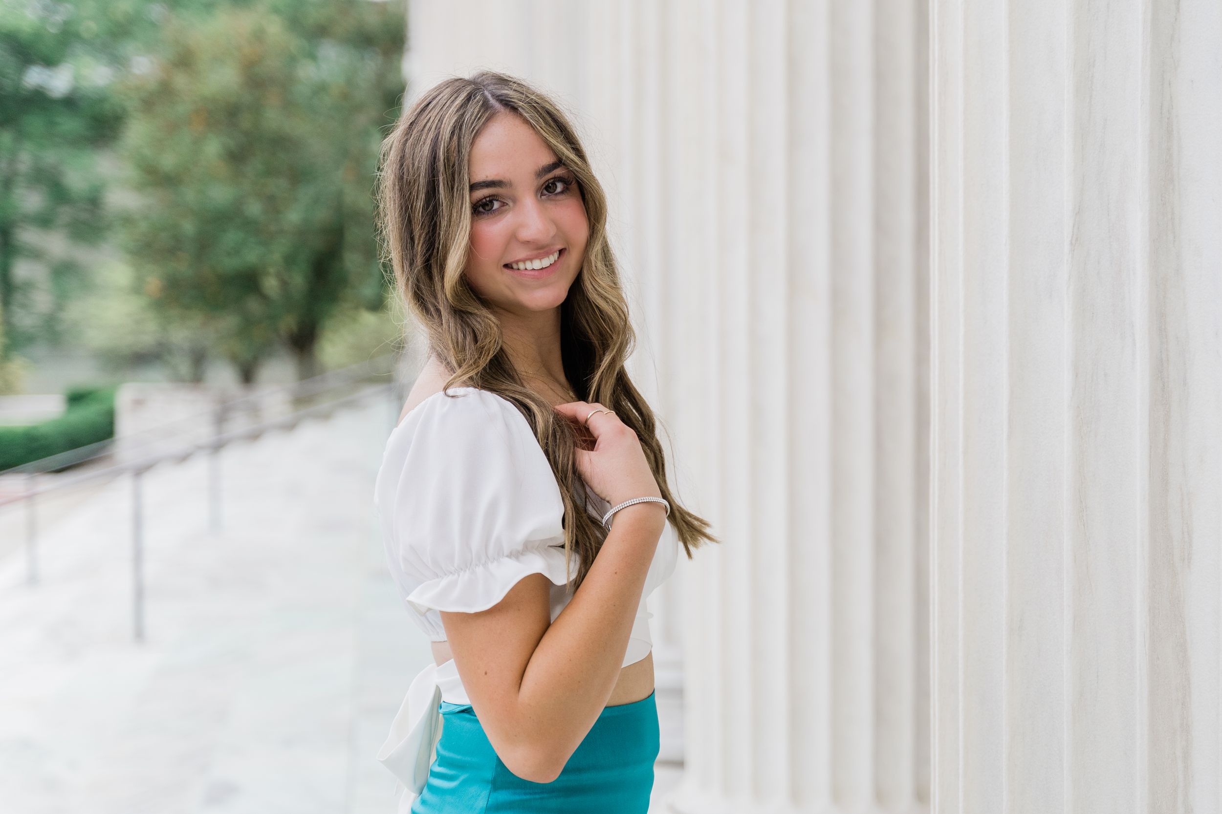 Someone poses in a white puff sleeve top and turquoise skirt against white columns outdoors.