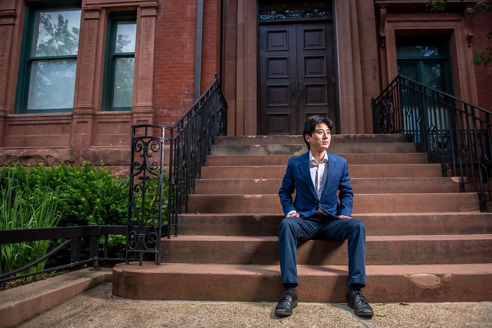 Someone in a navy blue suit sits on brownstone steps of a classic Brooklyn townhouse with iron railings and greenery.