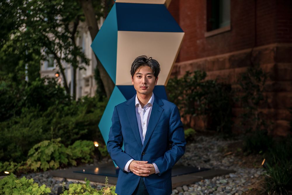 Person in a navy blue suit stands confidently outdoors against a brick building with geometric blue artwork.
