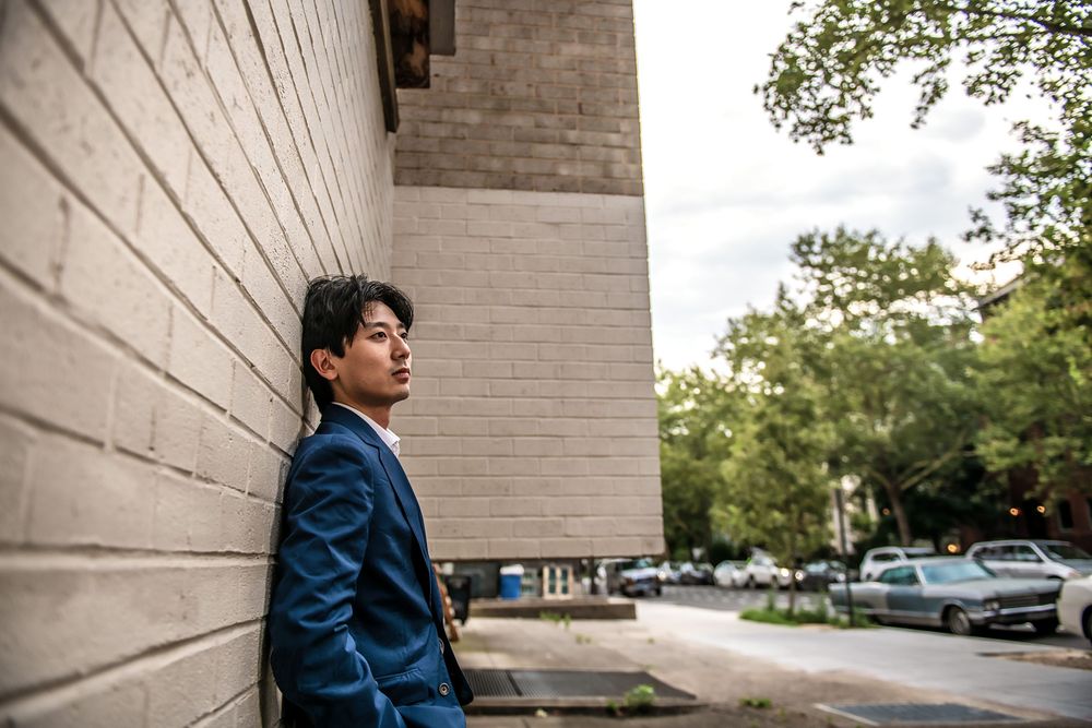 Someone leans against a beige brick wall wearing a blue denim jacket and black pants on an autumn day.