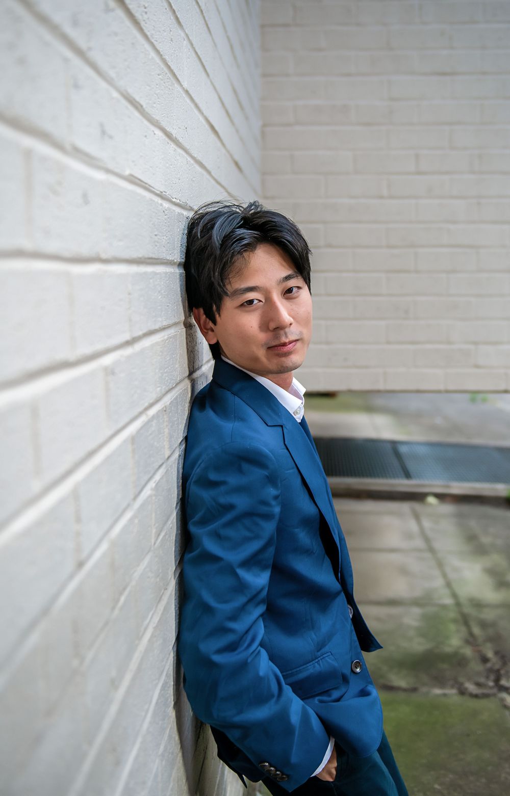 A person in a navy blue business suit leans against a white brick wall in a professional outdoor portrait.