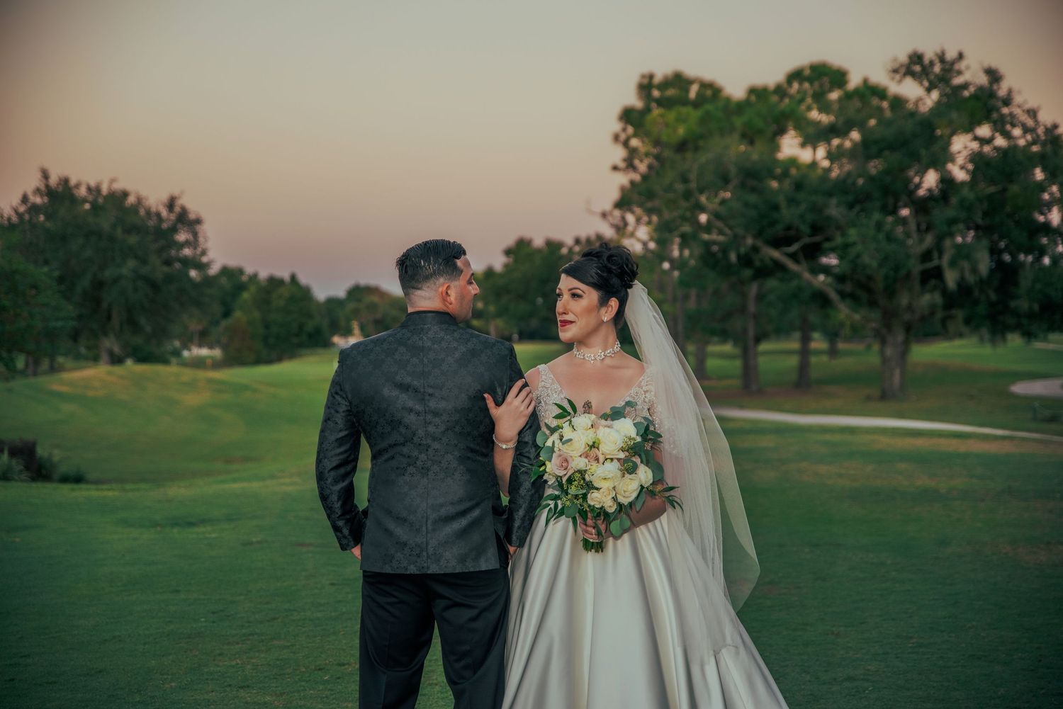 Bride and groom share romantic moment on golf course at dusk with bouquet.