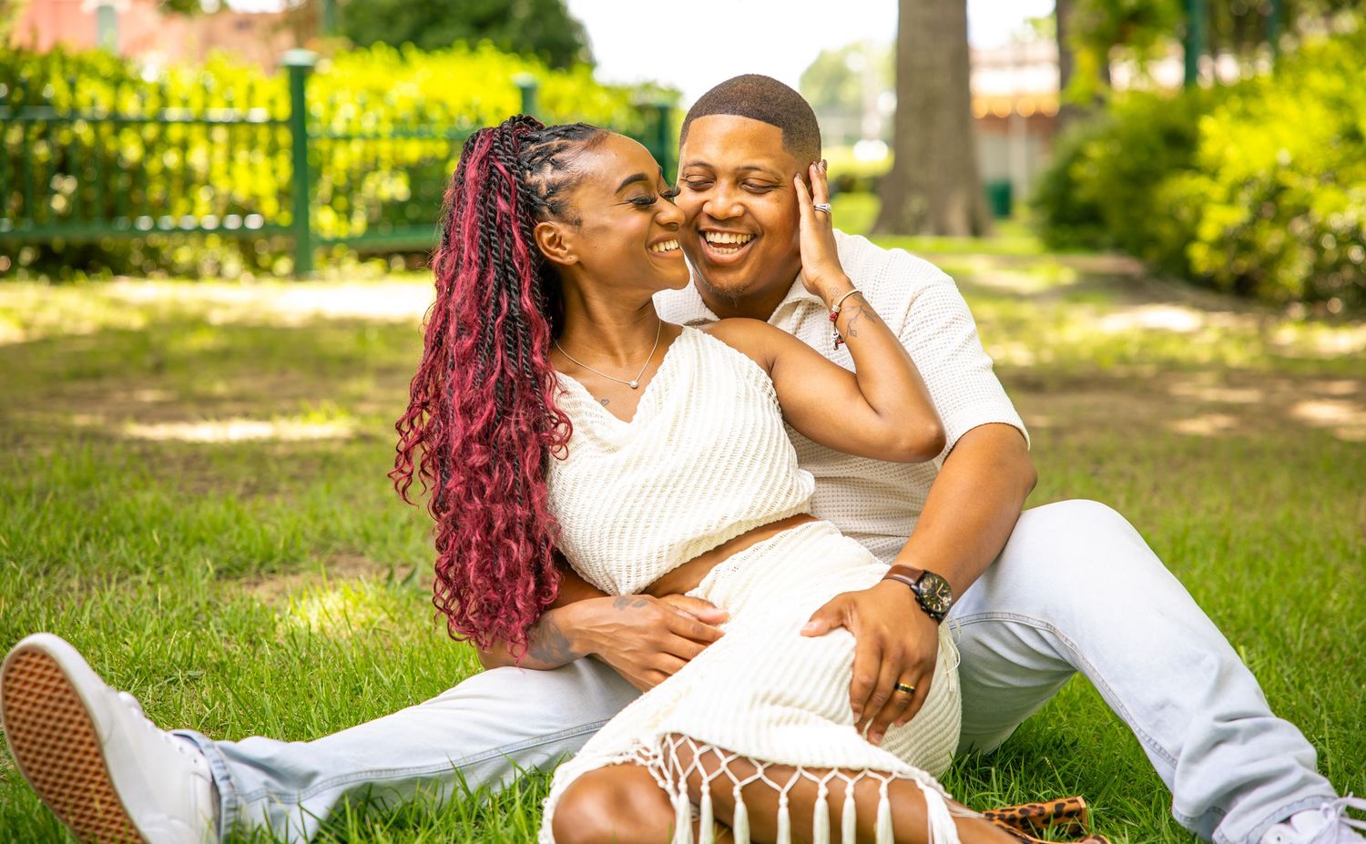 Couple in white outfits relaxing on green grass in a sun-lit park during an outdoor photo session.
