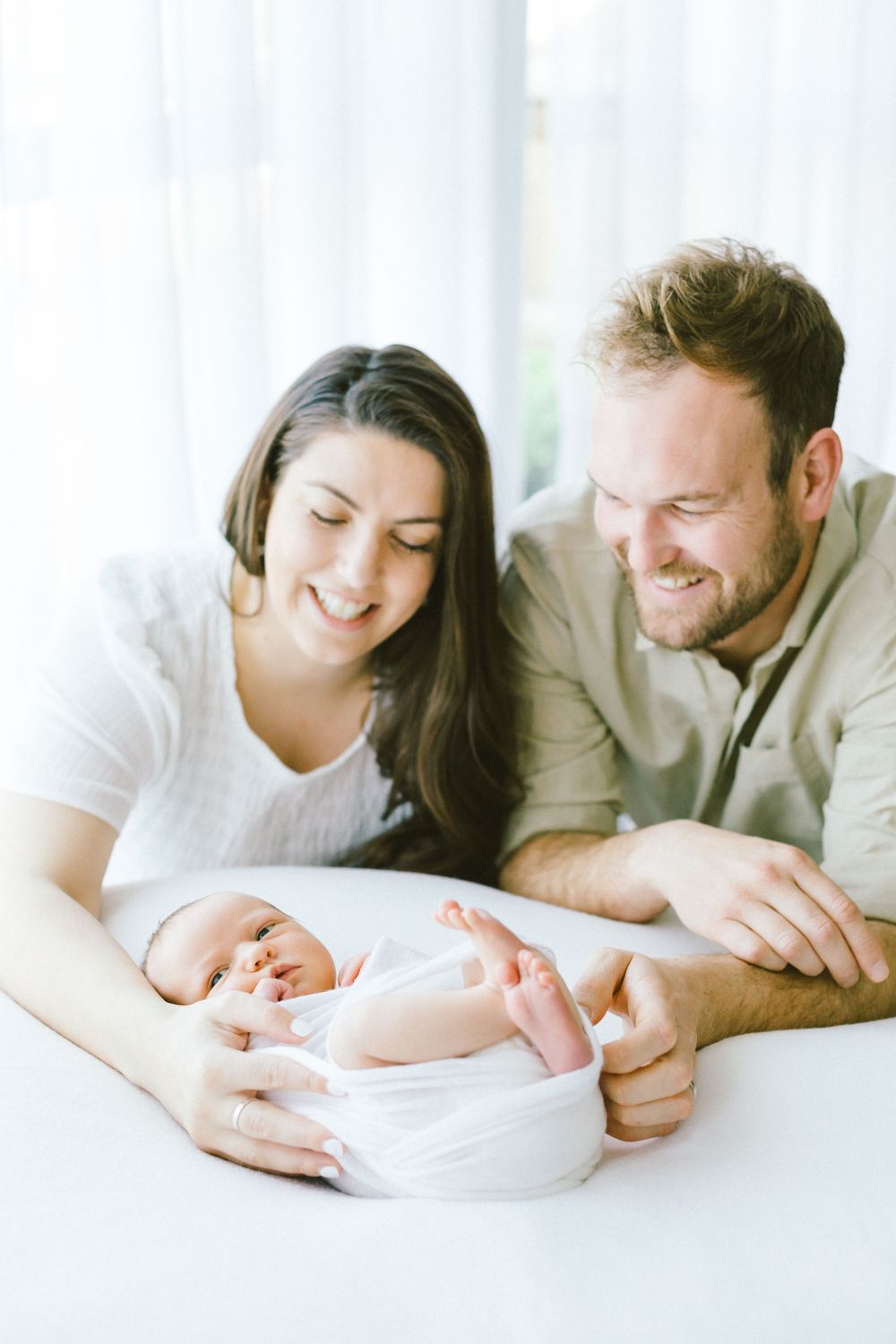 Parents smiling while laying on white surface with their newborn baby.