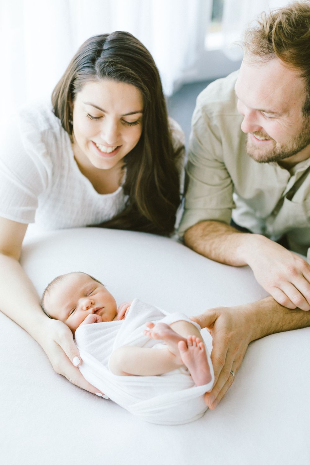 Parents gazing lovingly at their sleeping newborn on white surface.