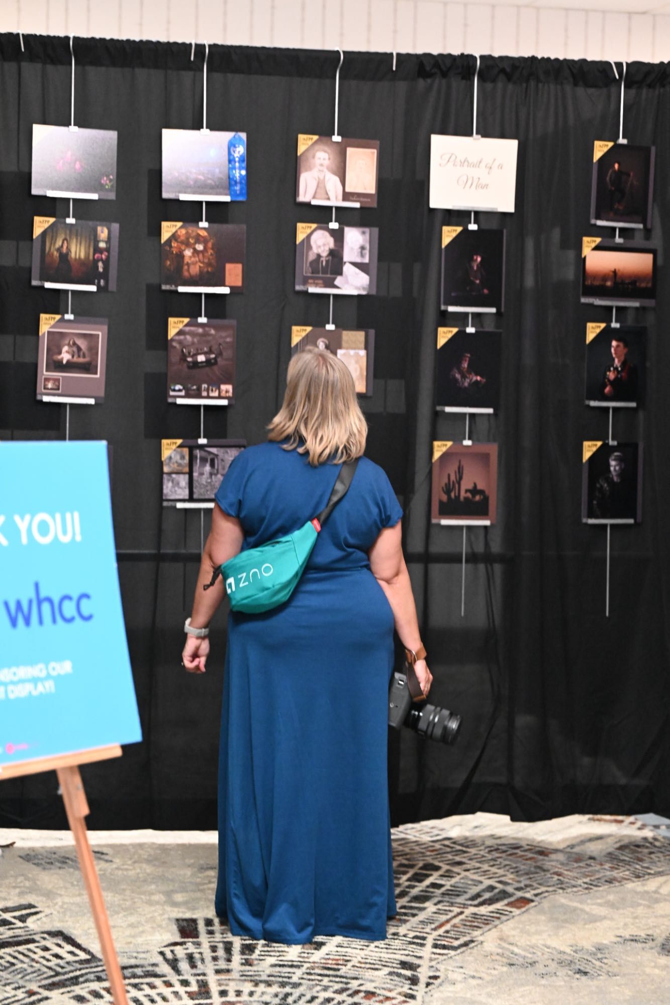 People in blue shirts view photographs displayed on dark walls with lighting at an art gallery exhibition.