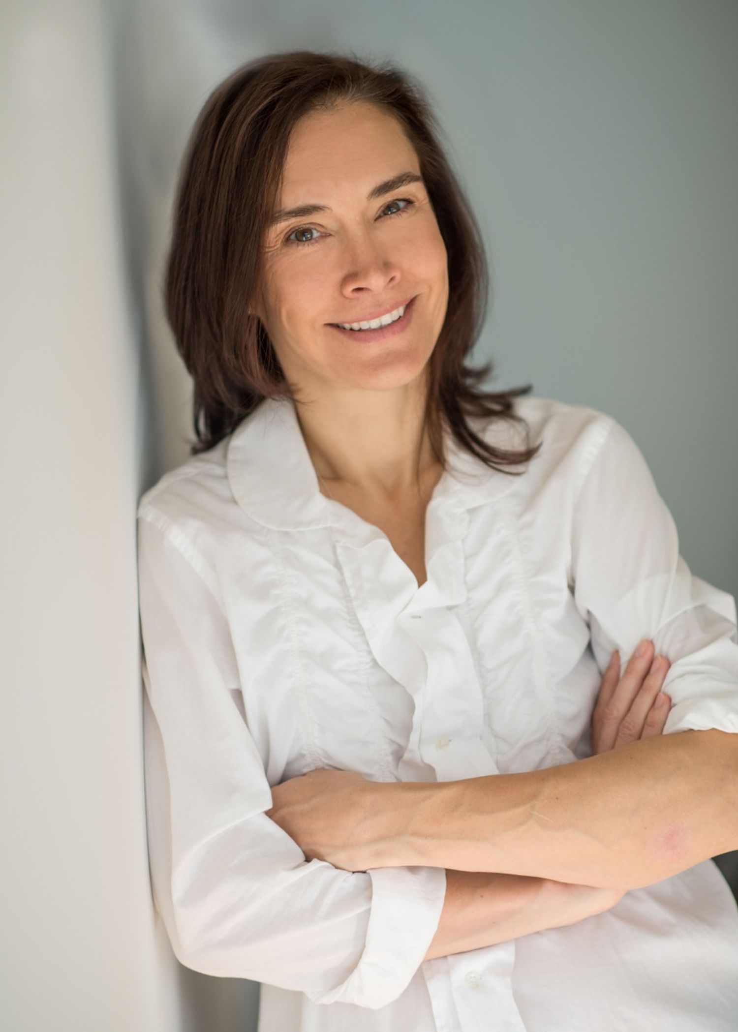 Professional woman in white button-down shirt smiling with arms crossed against light background.