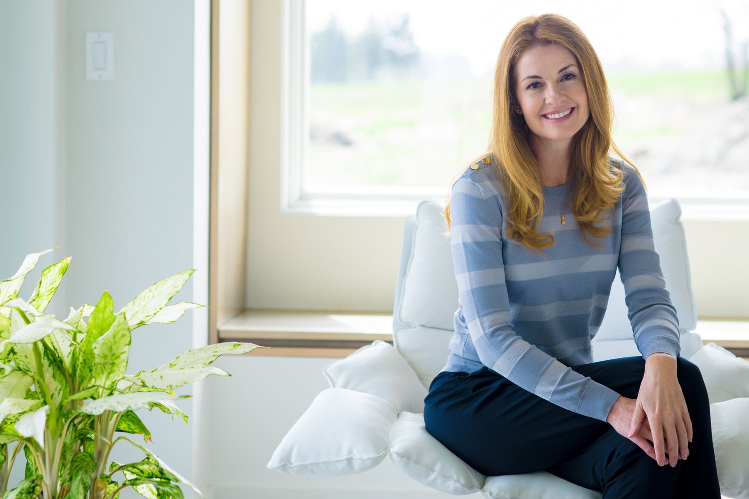 Woman in striped sweater relaxes on white couch near window with houseplant.