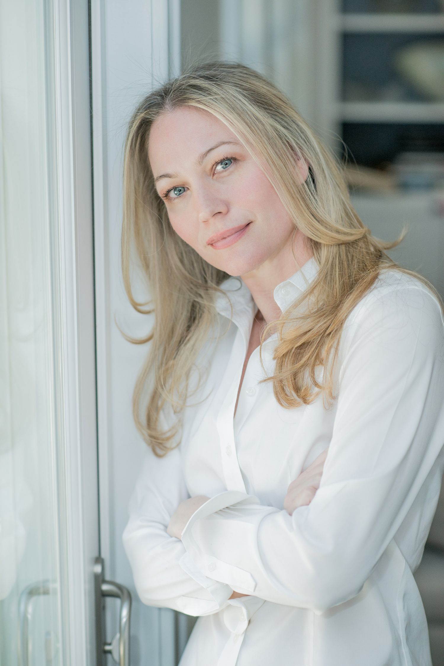 Portrait of a woman in a white button-up shirt standing near a window with her arms crossed.