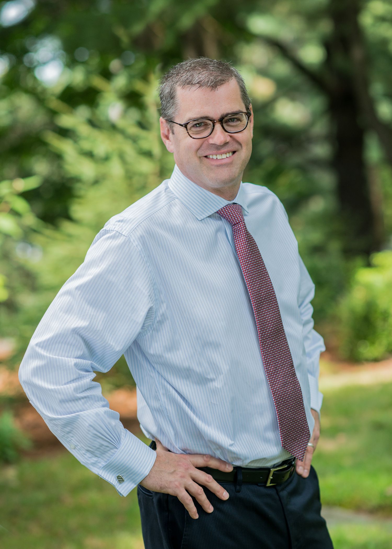 Professional portrait of someone in a light blue dress shirt and burgundy tie smiling outdoors with trees in background.