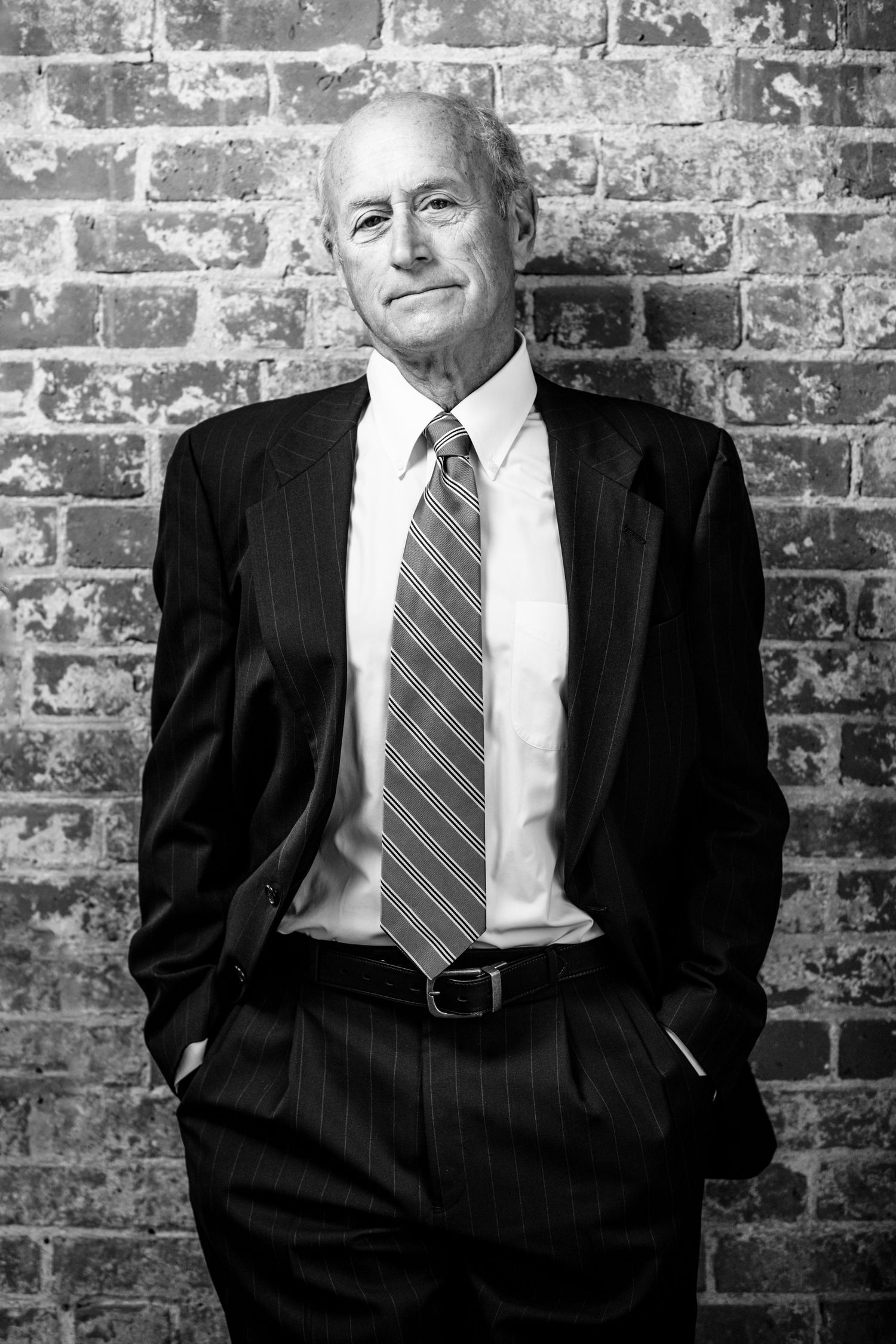 Black and white portrait of a senior businessman in a pinstripe suit and tie standing against a brick wall.