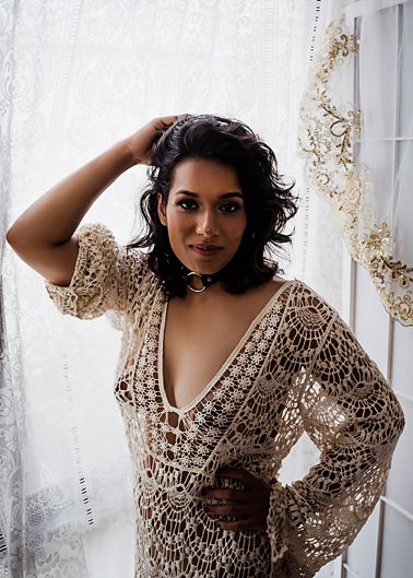 A woman in a crocheted beige lace dress poses near a window with flowing curtains in a romantic boudoir setting.